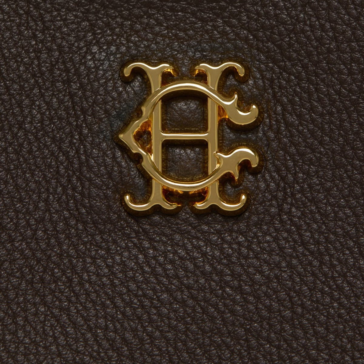 A close-up of a textured dark brown leather surface featuring an ornate gold metal monogram logo in the center.