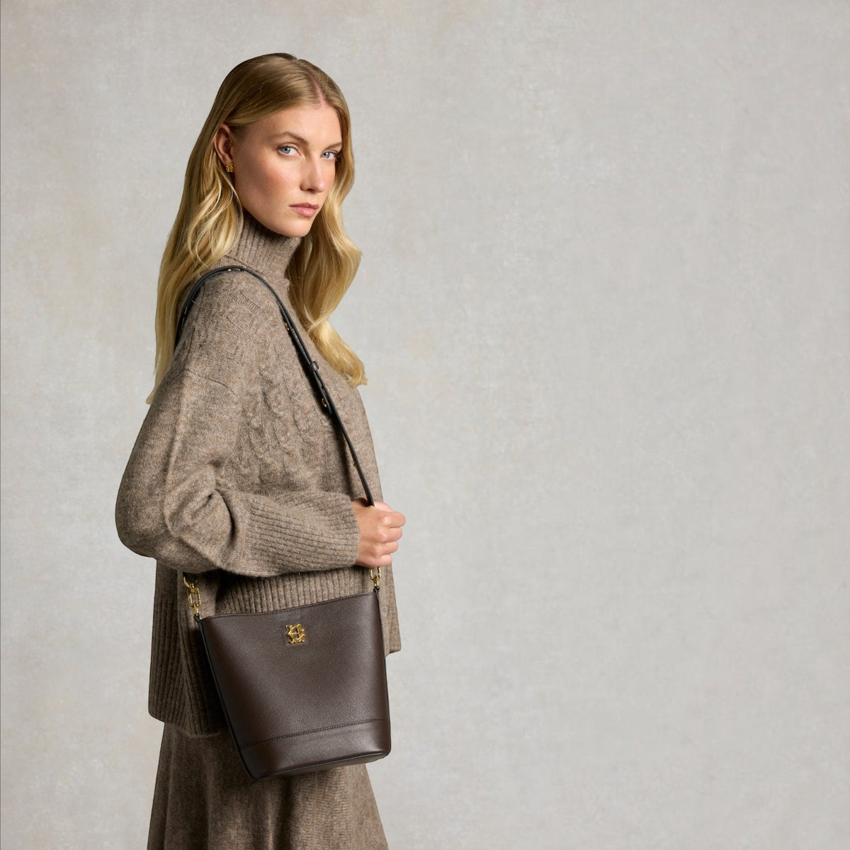 A blonde woman in a beige cable-knit turtleneck sweater and matching skirt carries a dark brown leather bucket bag with a gold logo clasp, posed against a textured light gray studio background.