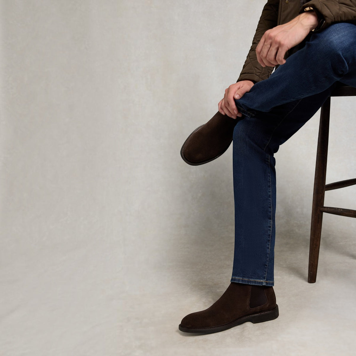 A man wearing dark brown suede Chelsea boots, blue jeans, and a brown jacket sits on a wooden stool against a textured light gray background.