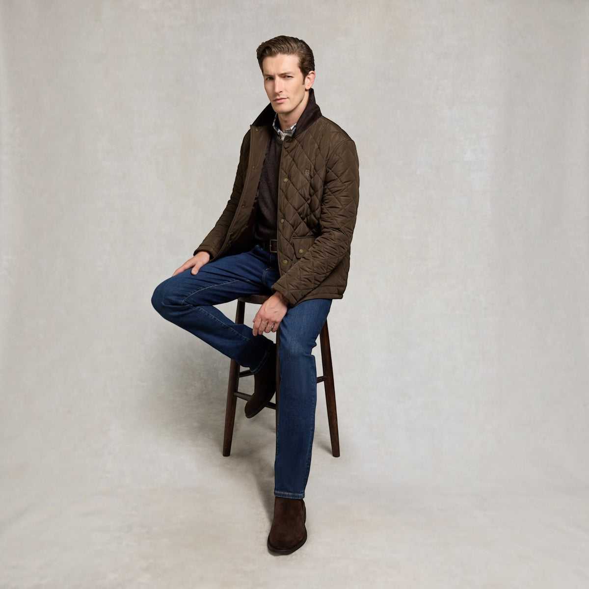 A man wearing a brown quilted jacket, blue jeans, and brown boots sits on a wooden stool against a textured light gray studio backdrop.