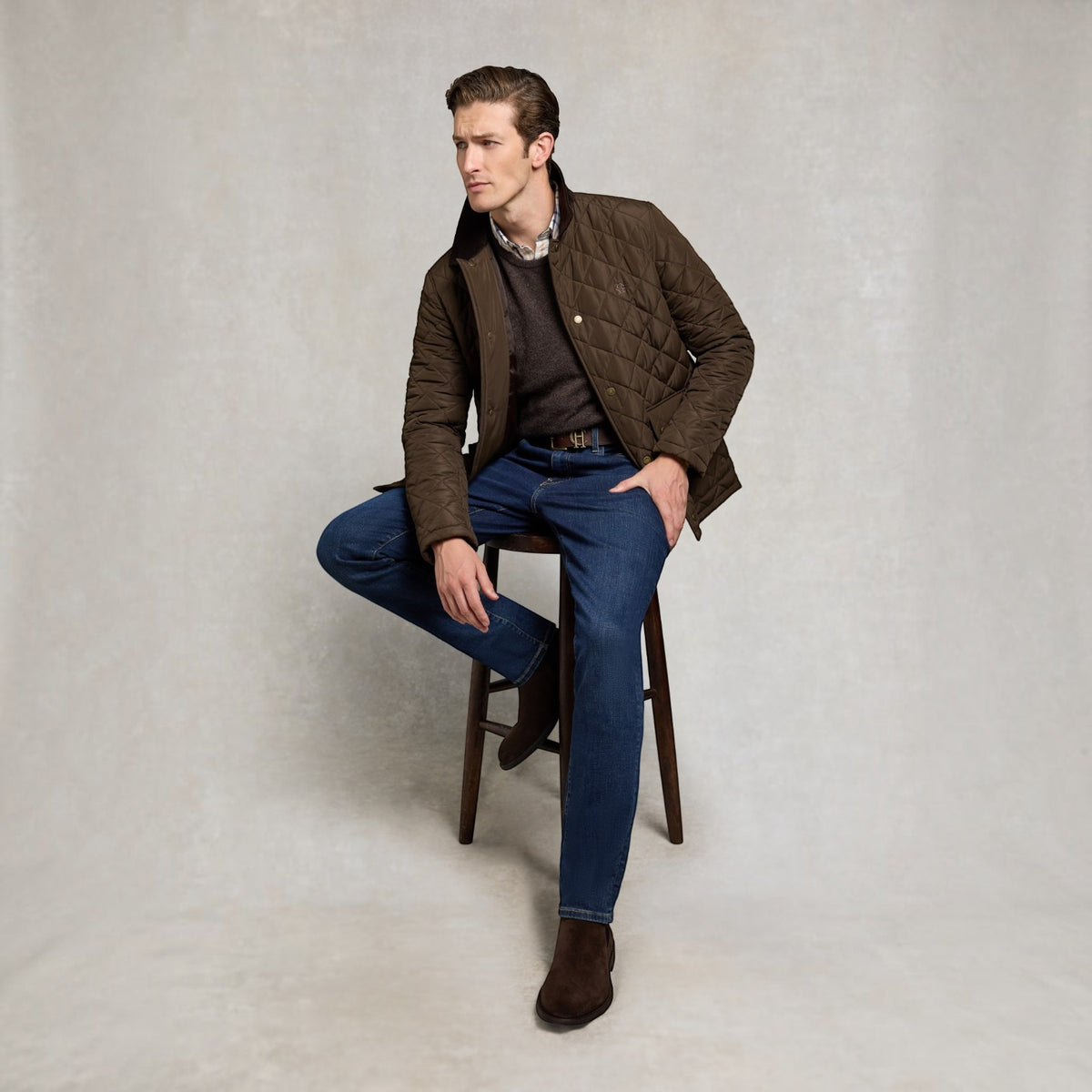 A man wearing a brown quilted jacket, dark brown sweater, blue jeans, and brown boots sits on a wooden stool against a neutral light gray studio background.