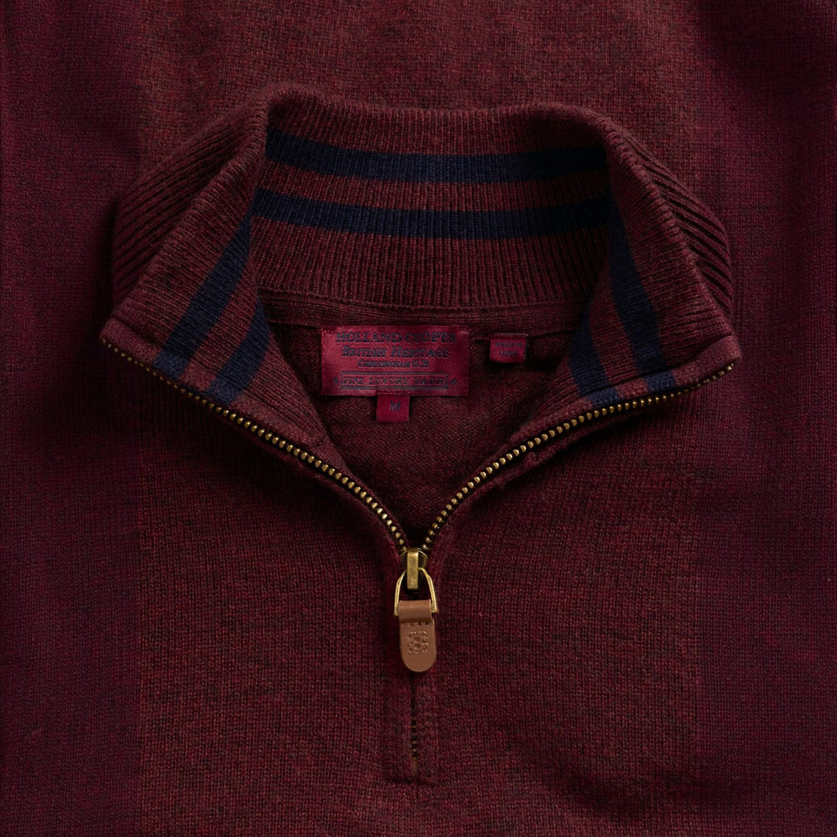 Close-up of a ribbed knit quarter-zip sweater in deep burgundy, featuring a stand collar with navy and burgundy stripes, a gold-tone zipper, and a visible red interior neck label.