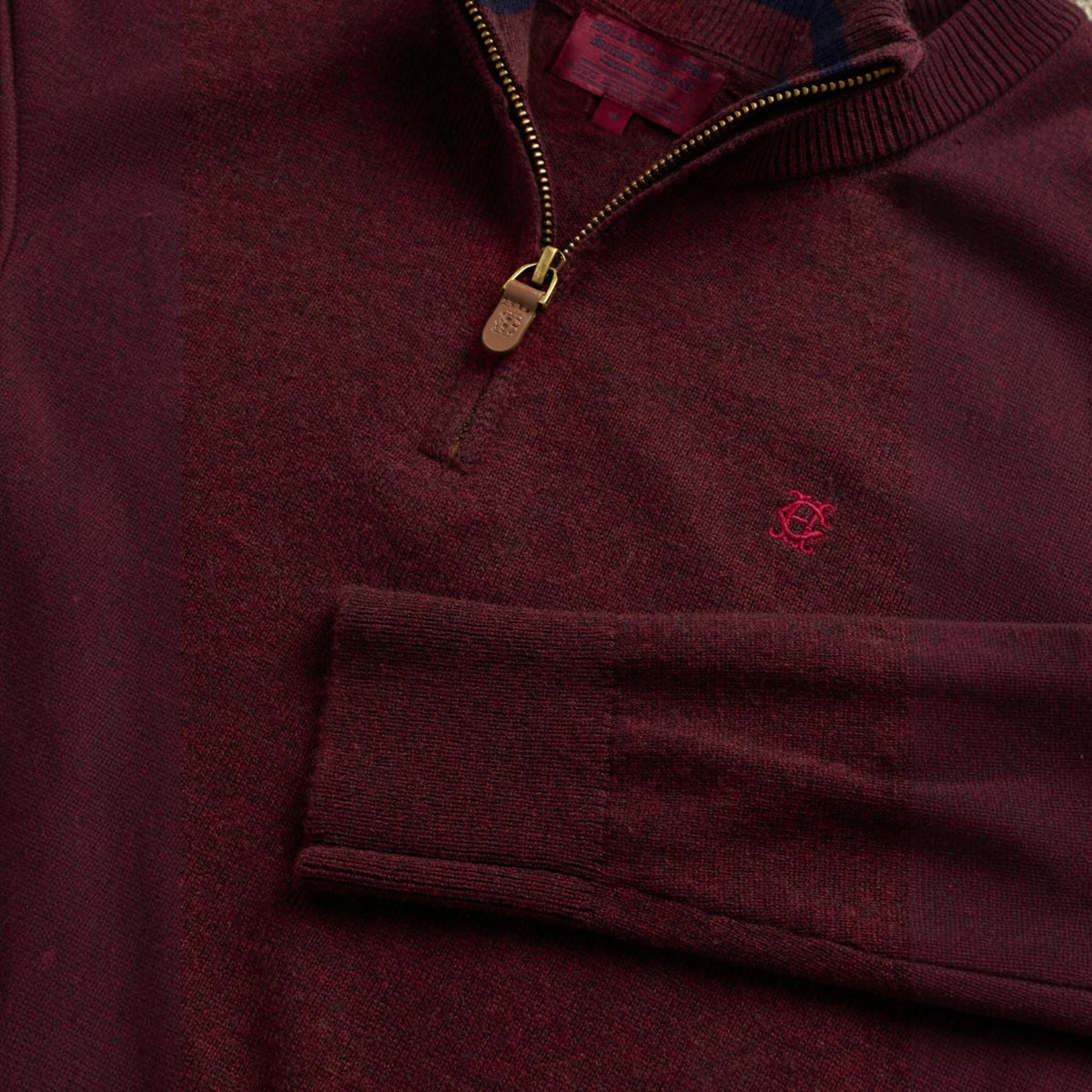 Close-up of a burgundy knit quarter-zip sweater with a small pink embroidered logo on the chest, a zippered pocket, and a gold-tone zipper pull.