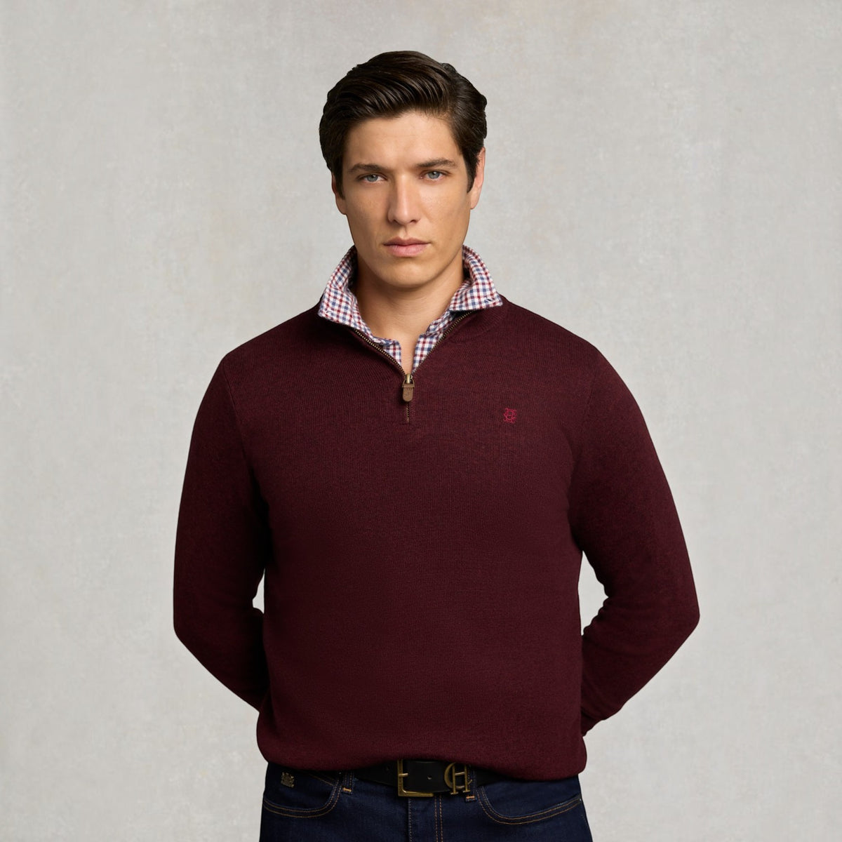 A man wearing a burgundy quarter-zip sweater over a red and white checkered collared shirt, paired with dark blue jeans, standing against a textured light gray background.