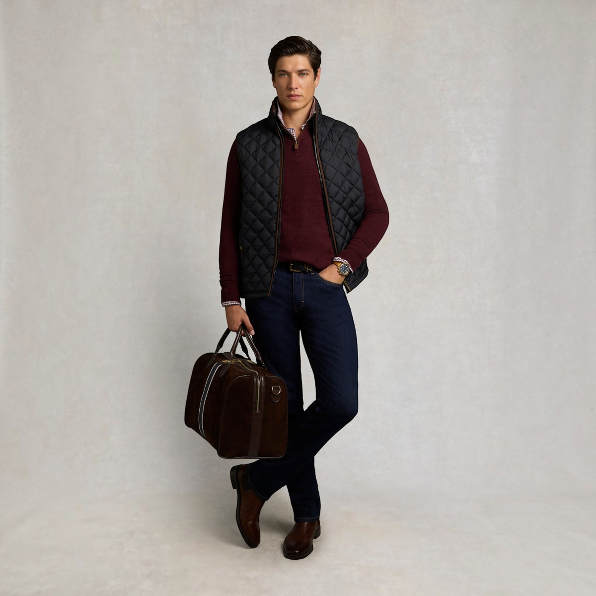 A man stands against a light gray studio background wearing a quilted black vest over a burgundy sweater, dark blue jeans, brown shoes, and holding a brown leather duffel bag.