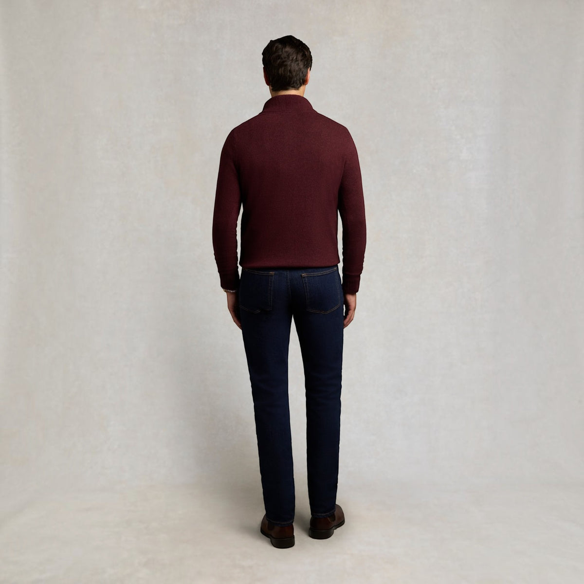 A man viewed from behind wearing a maroon mock-neck knit sweater and dark blue jeans, standing against a light textured studio backdrop.
