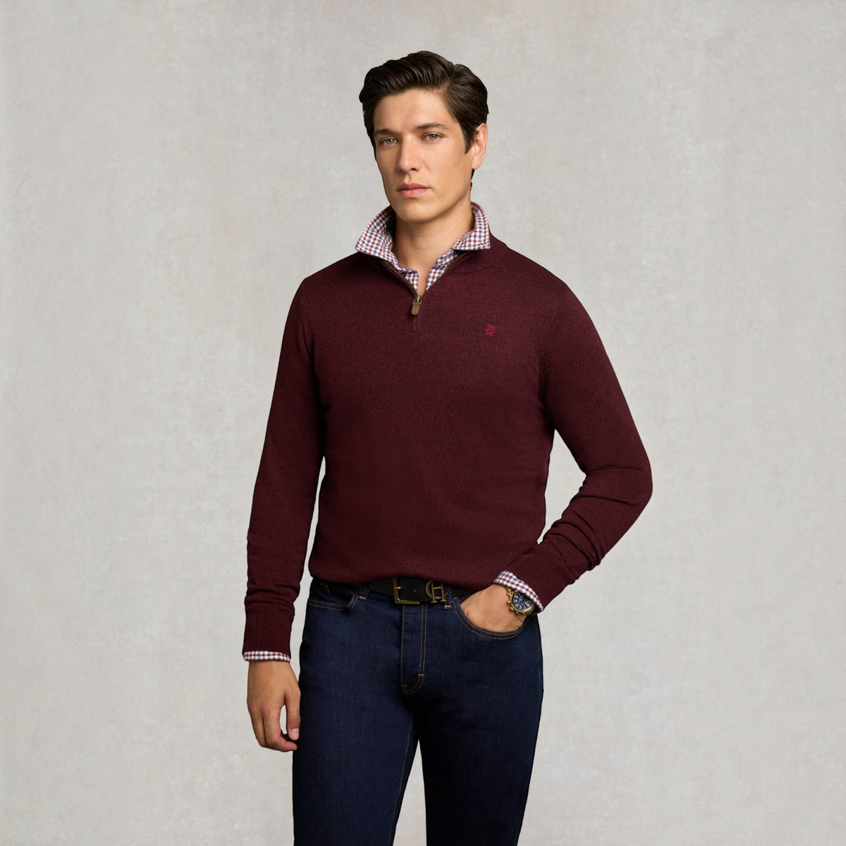 A man wearing a burgundy quarter-zip knit sweater over a red and white checkered collared shirt, paired with dark blue jeans, standing against a textured light gray background.
