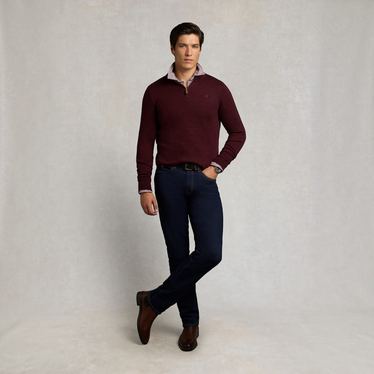 A man modeling a burgundy quarter-zip knit sweater over a patterned collared shirt, paired with dark blue jeans and brown leather shoes, against a neutral light gray studio backdrop.