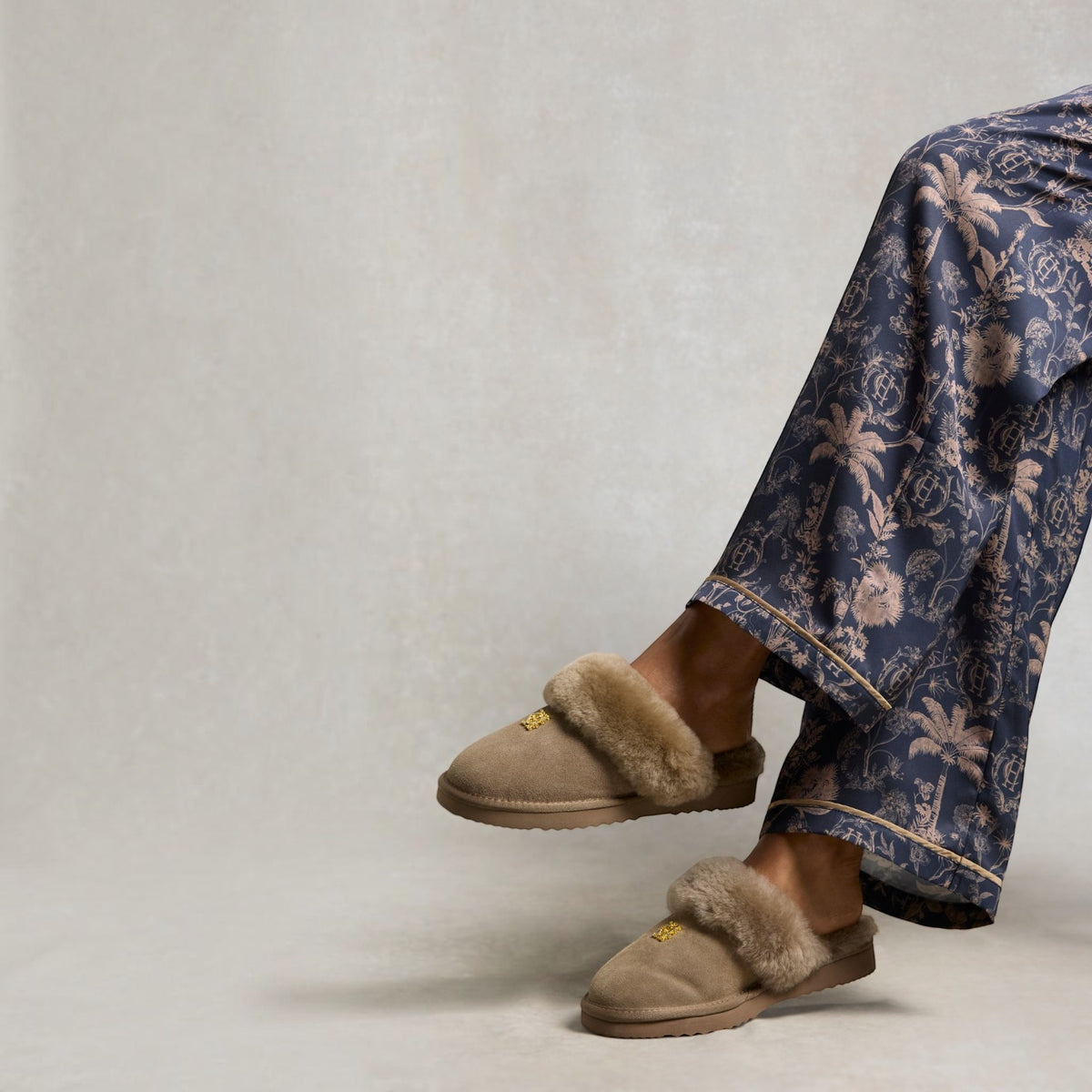 A person wearing beige slip-on slippers with fluffy trim and navy floral pajama pants, photographed against a light neutral studio background.