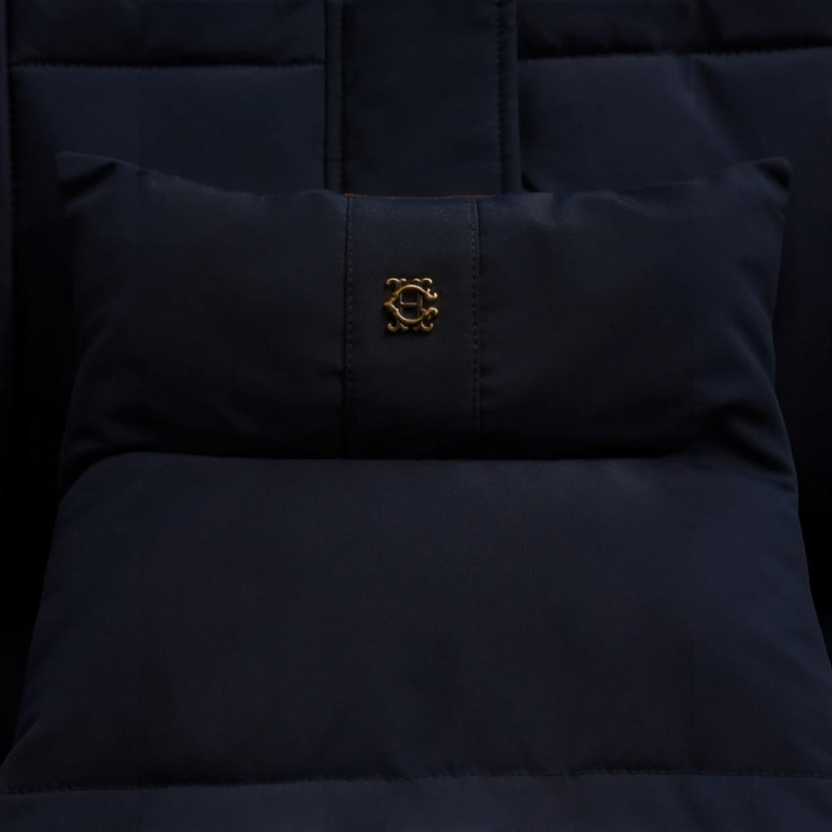 A close-up of a dark navy quilted cushion or pillow with a small gold embroidered monogram logo in the center, set against matching dark navy fabric.