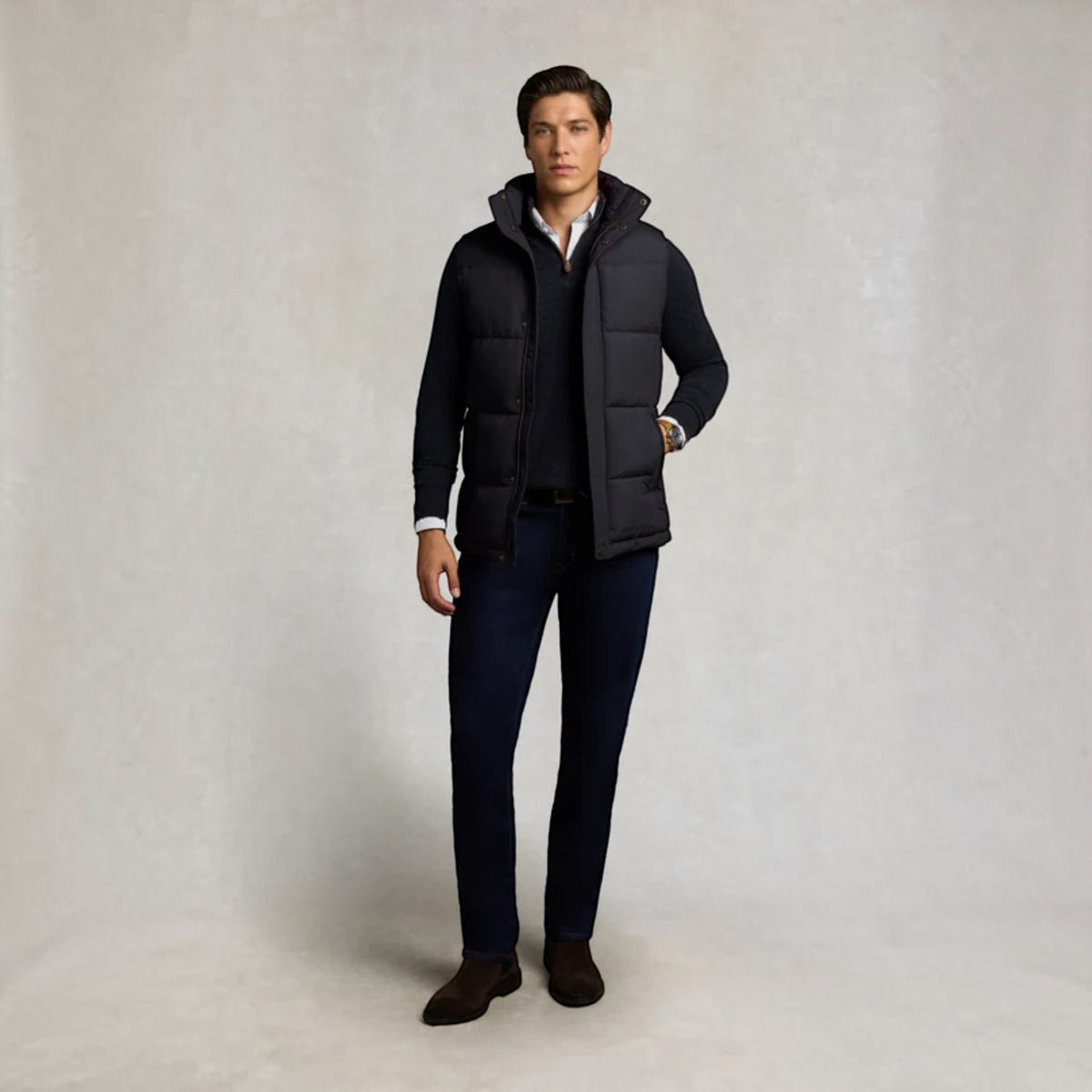 A man stands against a light gray studio backdrop wearing a dark navy puffer vest over a dark sweater and collared shirt, with dark trousers and brown shoes.