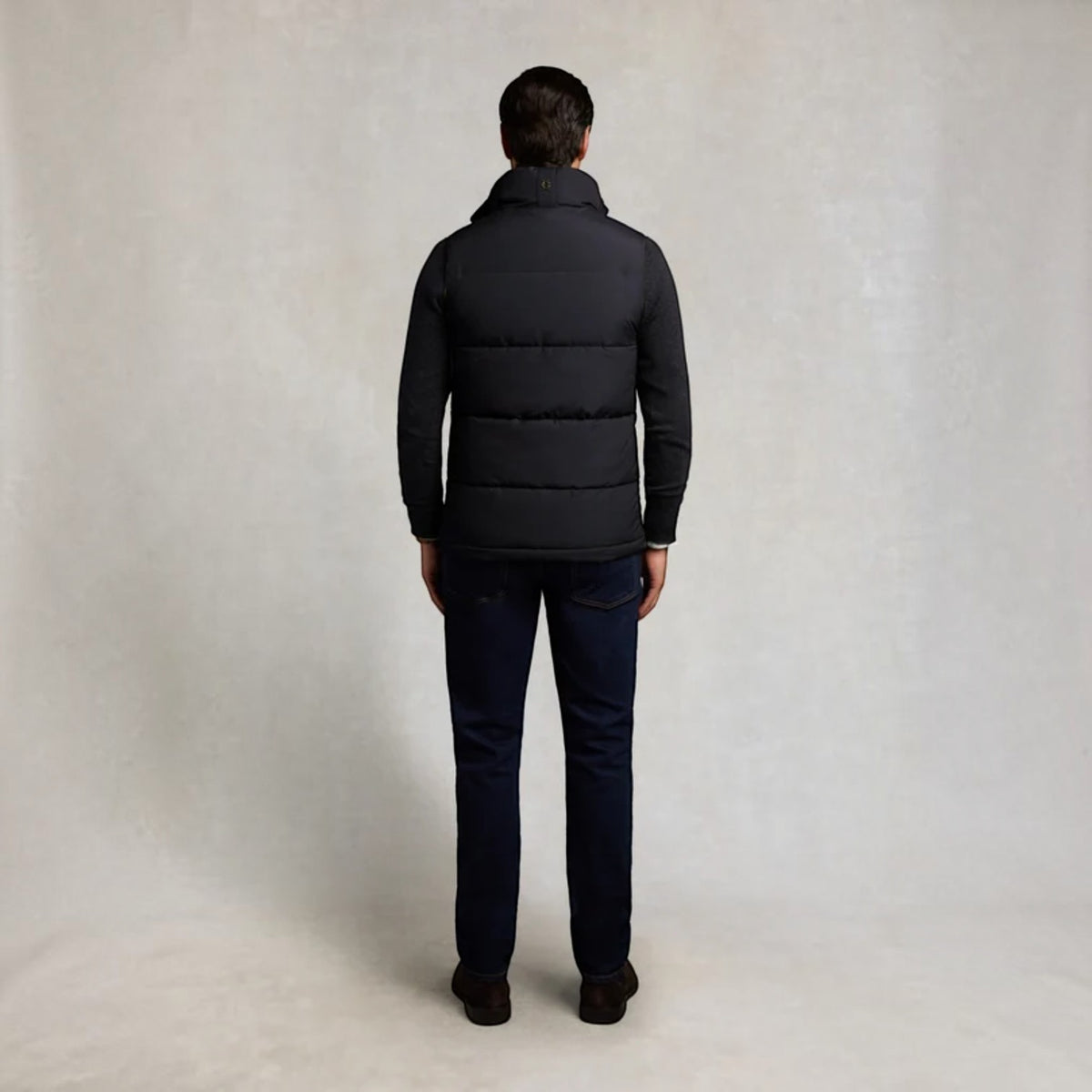 A man viewed from behind wearing a black puffer jacket, dark navy pants, and brown shoes, standing against a neutral light gray studio backdrop.