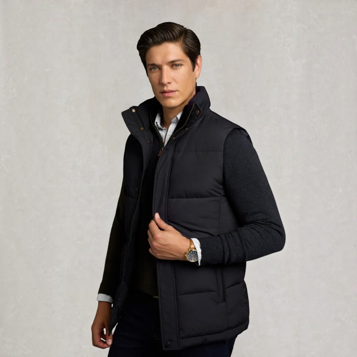 A man wearing a dark navy puffer vest over a black jacket and white collared shirt, standing against a textured light gray background.