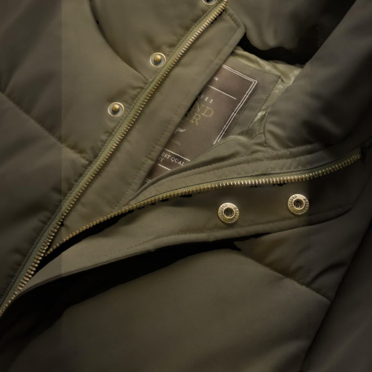 Close-up of a dark olive green puffer jacket showing the inner collar, a gold-tone zipper, snap buttons, and a brand label.