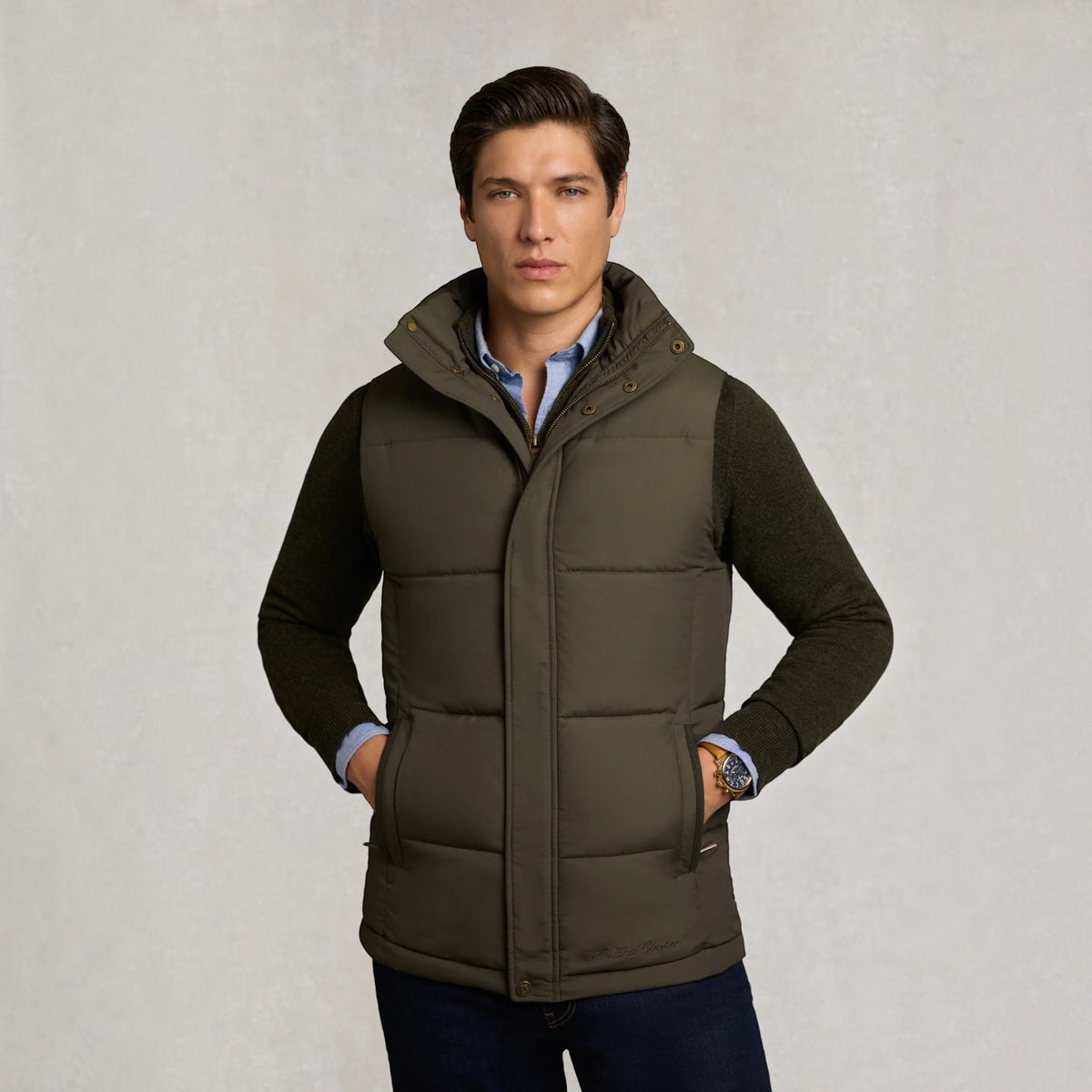 A man wearing an olive green quilted puffer vest over a dark green sweater, light blue collared shirt, and dark jeans, standing against a textured light gray background.