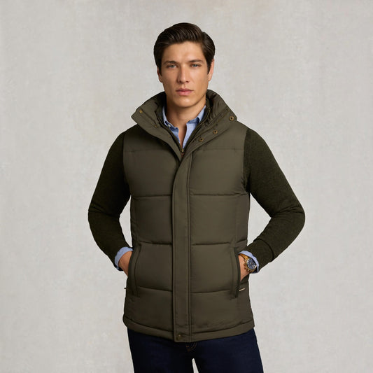 A man wearing an olive green quilted puffer vest over a dark green sweater, light blue collared shirt, and dark jeans, standing against a textured light gray background.