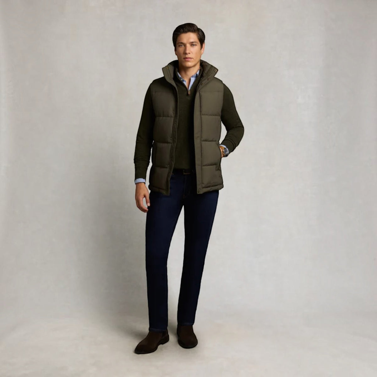 A man modeling a dark olive green quilted puffer vest over a dark green sweater, a light blue collared shirt, dark blue jeans, and brown shoes, standing against a neutral light gray studio backdrop.