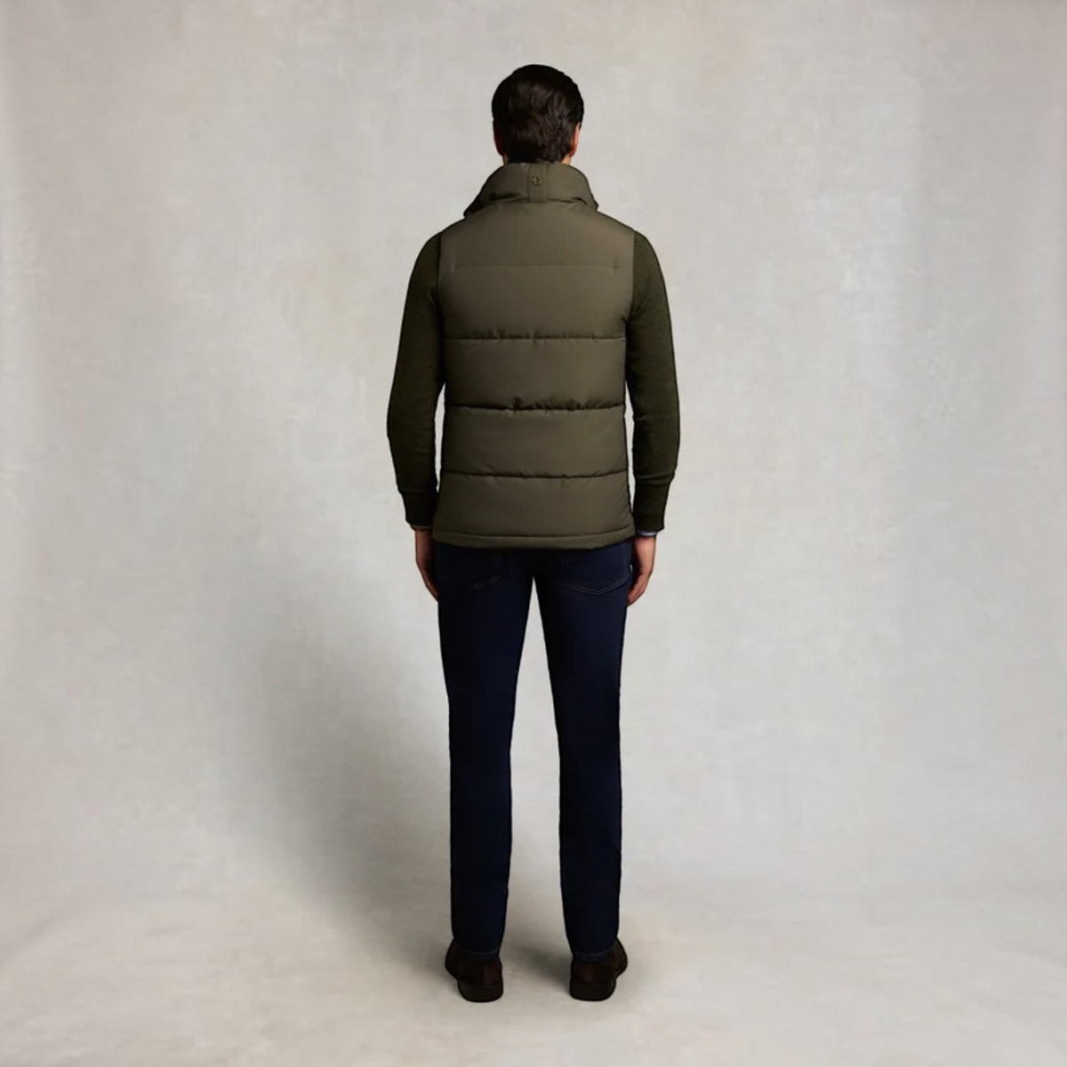 A man viewed from behind wearing an olive green puffer vest over a dark long-sleeve shirt, paired with dark navy pants and brown shoes, standing against a light gray textured studio backdrop.
