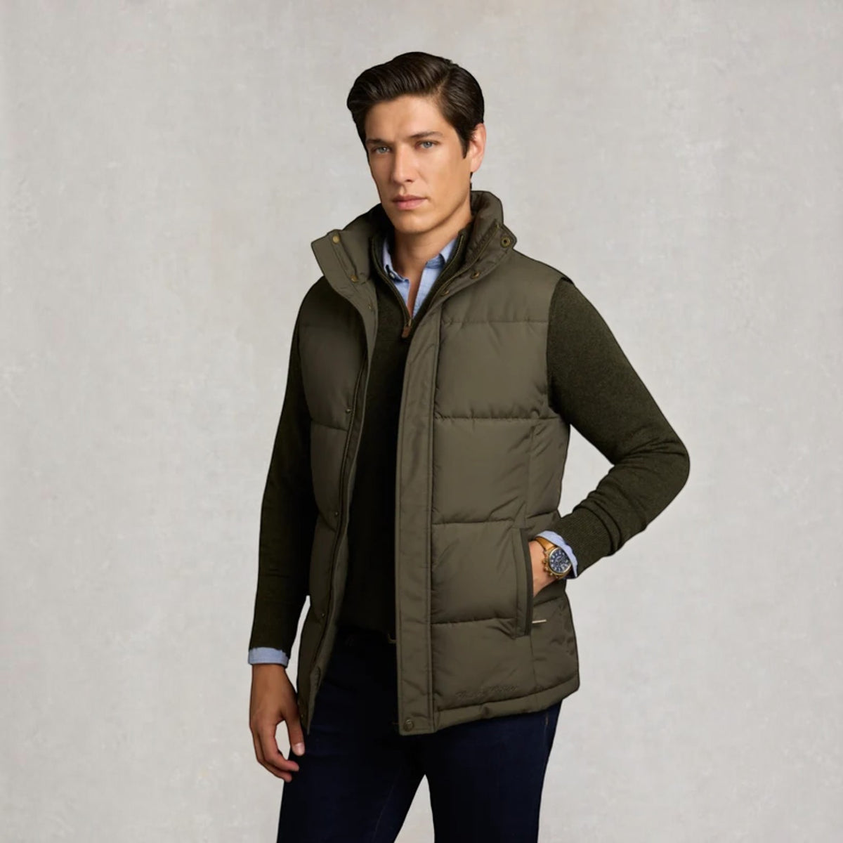 A man wearing an olive green puffer vest over a dark sweater and light blue collared shirt, standing against a textured light gray studio background.