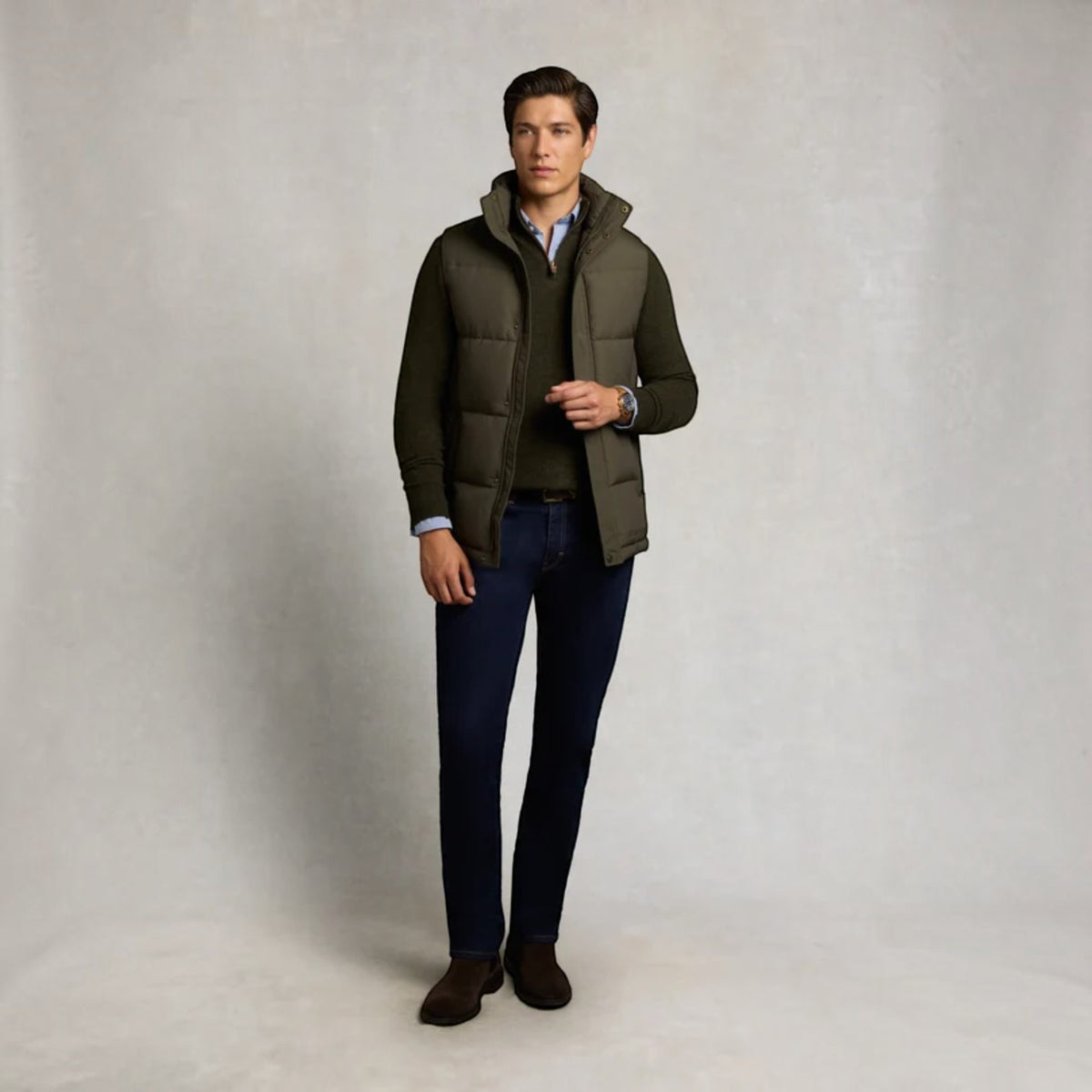 A man modeling an olive green hooded puffer vest over a dark sweater and light blue shirt, paired with dark blue jeans and brown shoes, against a neutral light gray studio backdrop.