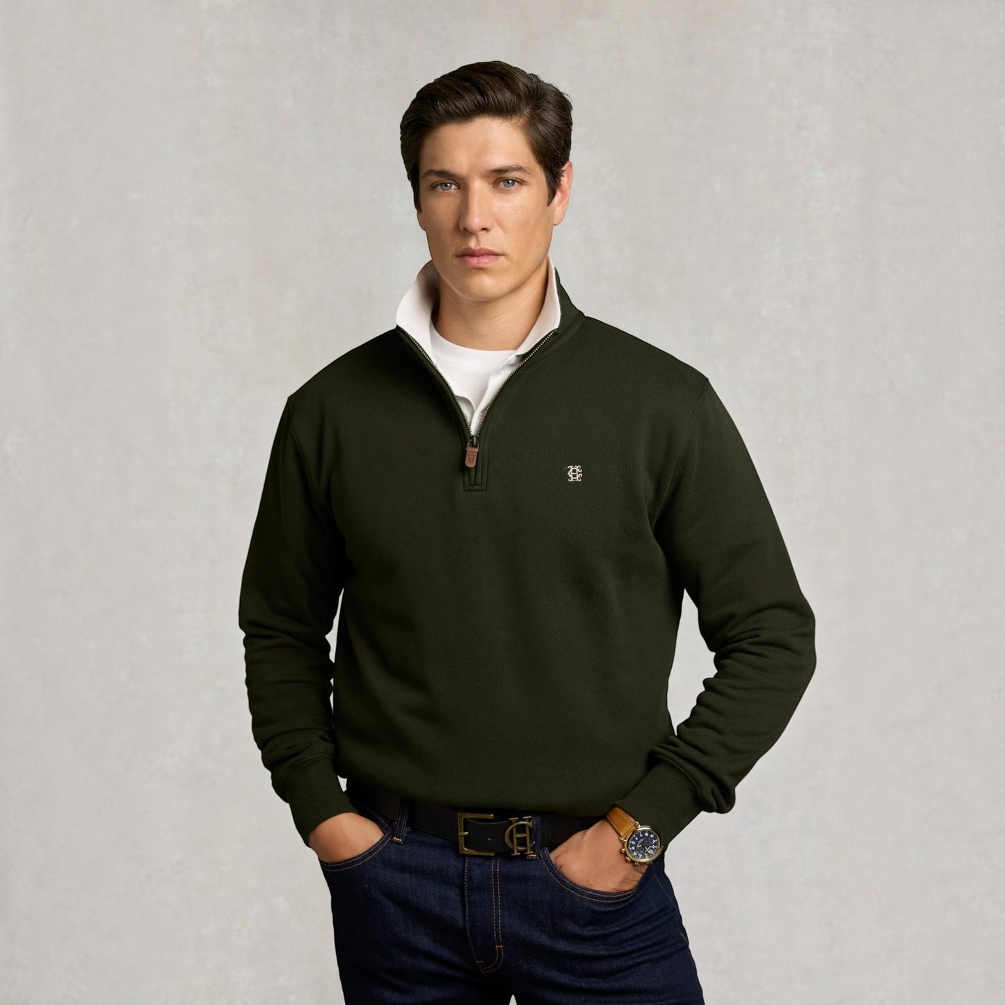 A man wearing an olive green quarter-zip pullover sweater over a white t-shirt and dark blue jeans, standing against a textured light gray background.