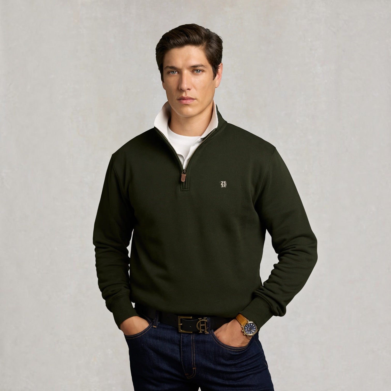 A man wearing an olive green quarter-zip pullover sweater over a white t-shirt and dark blue jeans, standing against a textured light gray background.