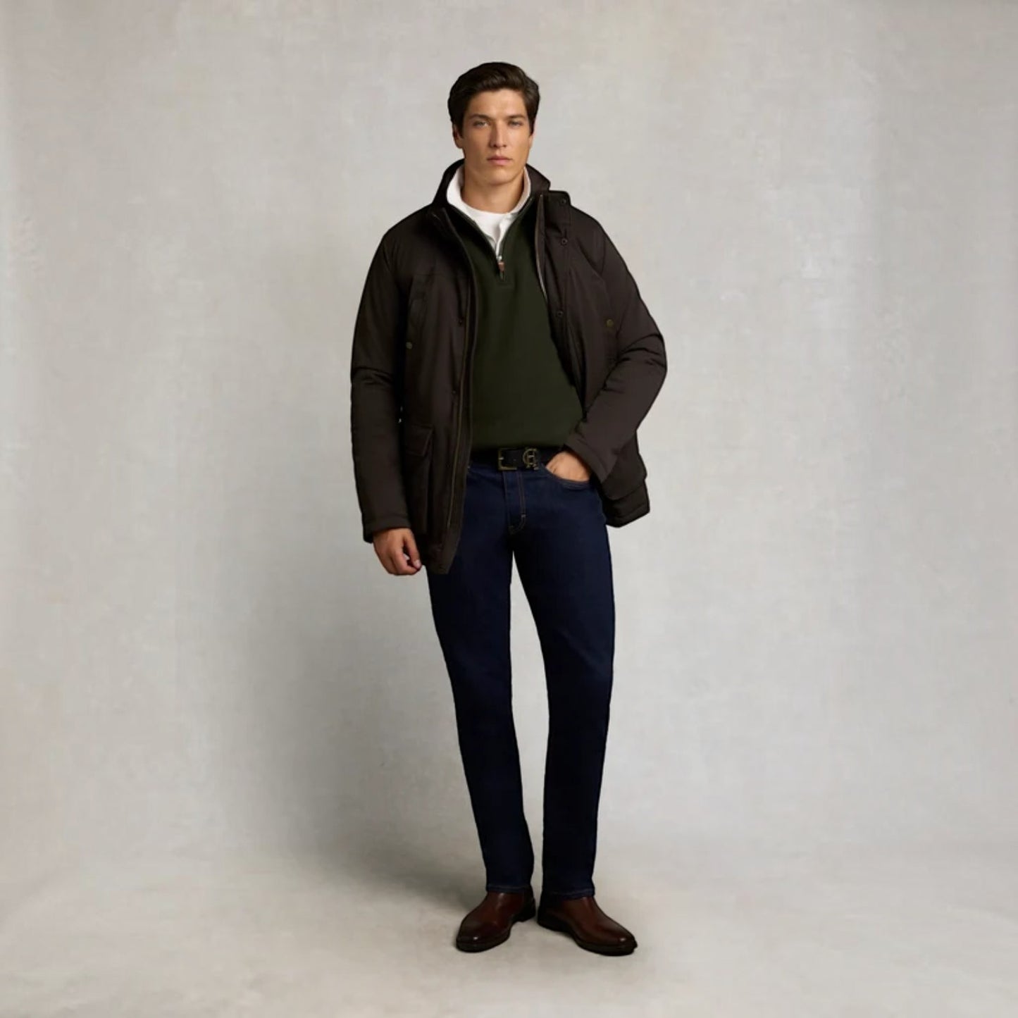 A man modeling a dark brown hooded jacket over a green quarter-zip sweater, dark blue jeans, and brown leather shoes, standing against a neutral light gray studio backdrop.
