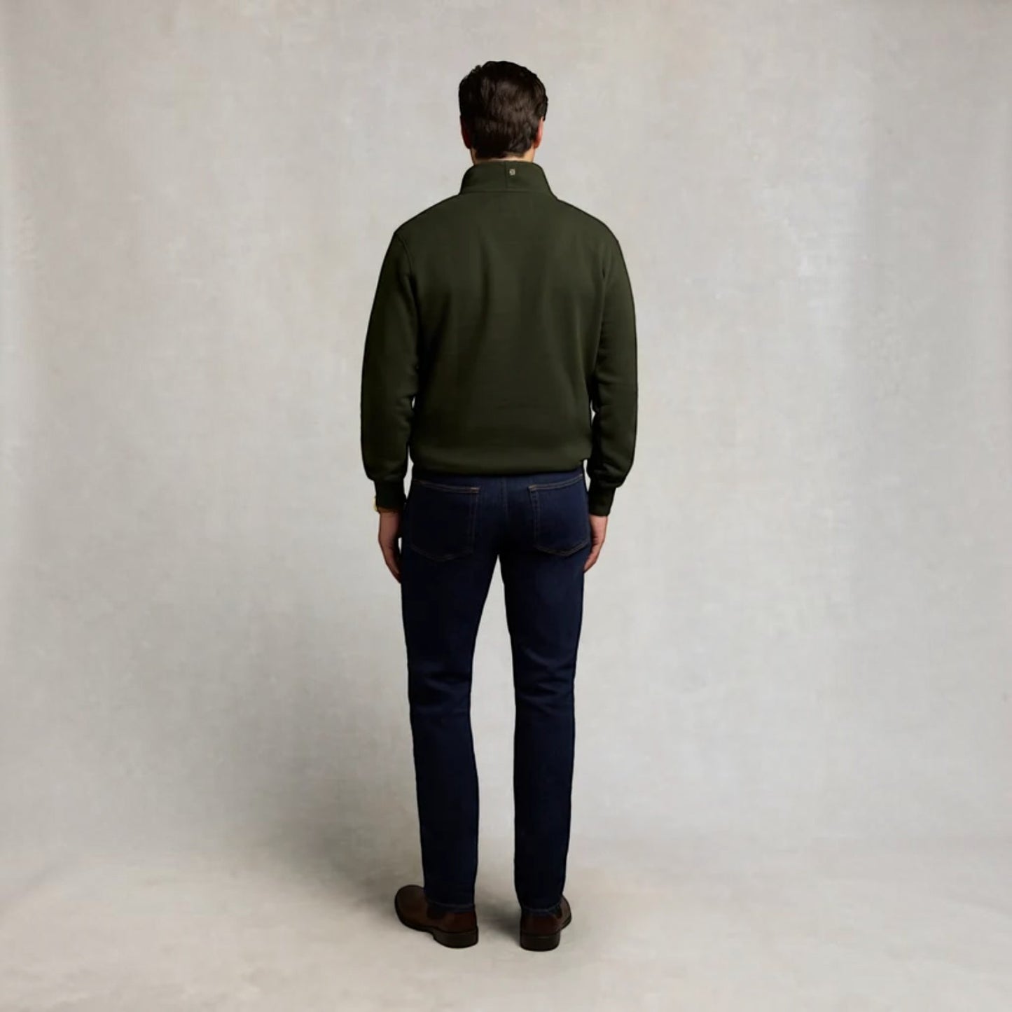 A man viewed from behind wearing an olive green quarter-zip fleece pullover, dark navy blue jeans, and brown leather shoes, standing against a neutral textured light gray backdrop.