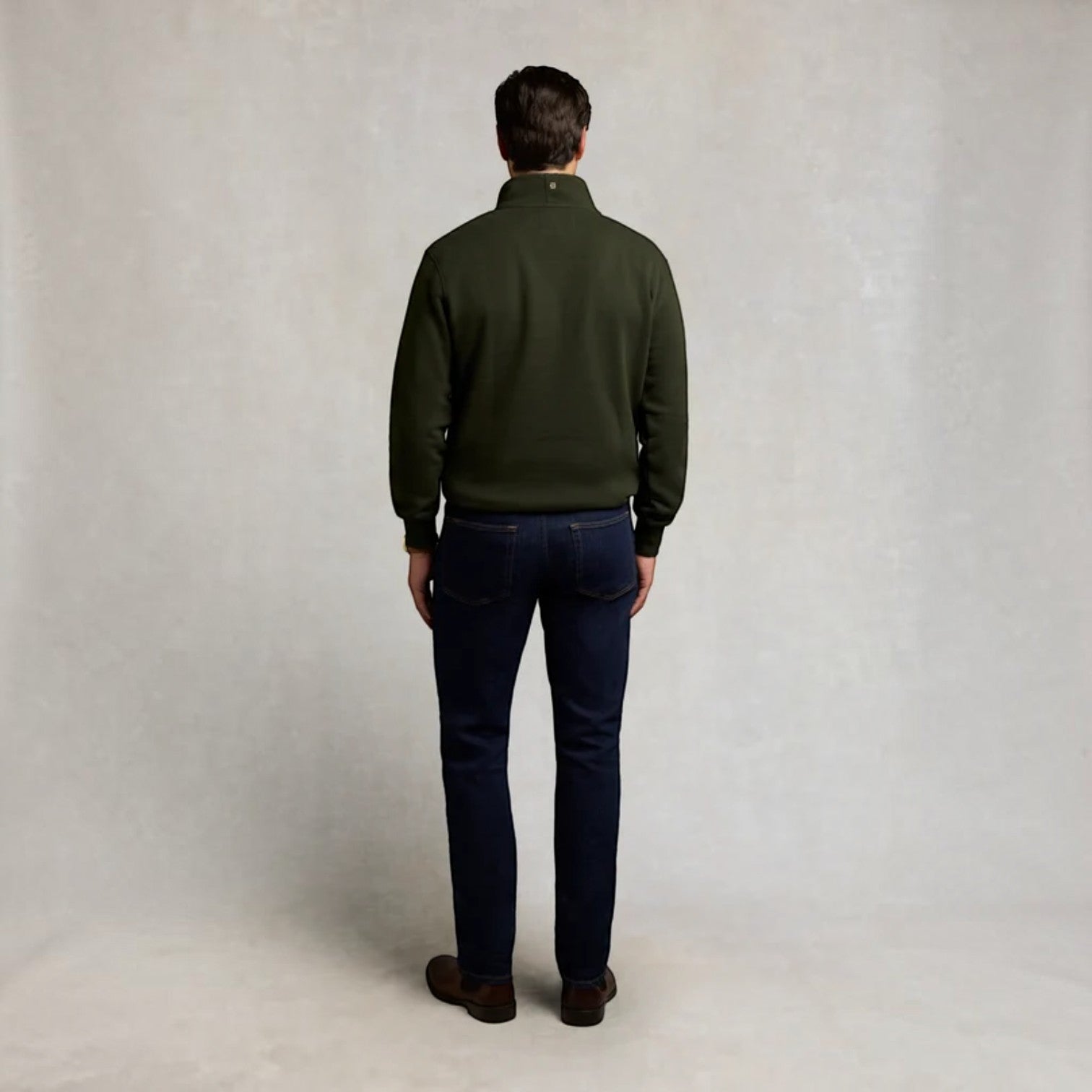 A man viewed from behind wearing an olive green quarter-zip fleece pullover, dark navy blue jeans, and brown leather shoes, standing against a neutral textured light gray backdrop.