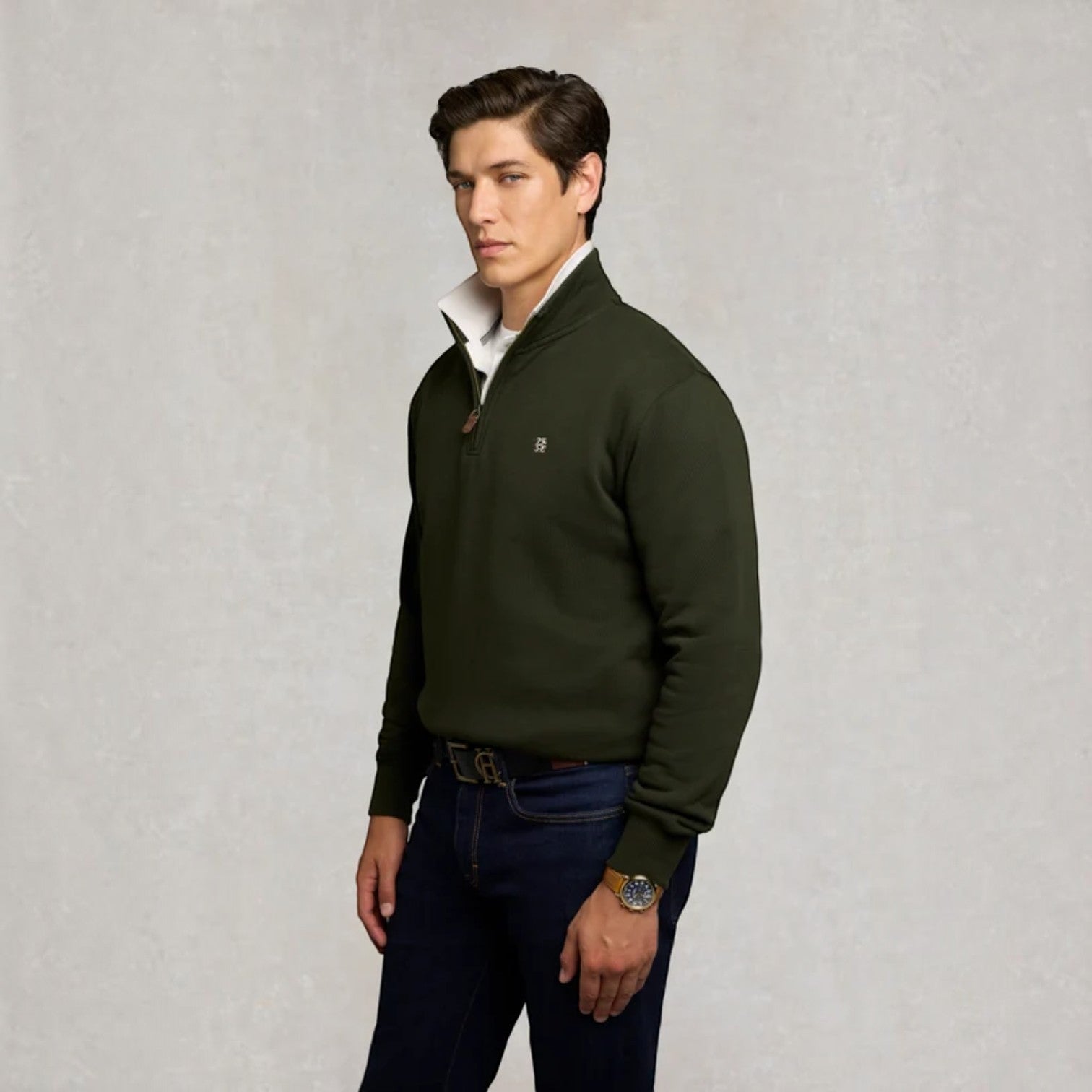 A man wearing a dark olive green quarter-zip pullover sweater with a white collar insert, paired with dark blue jeans, standing against a textured light gray background.