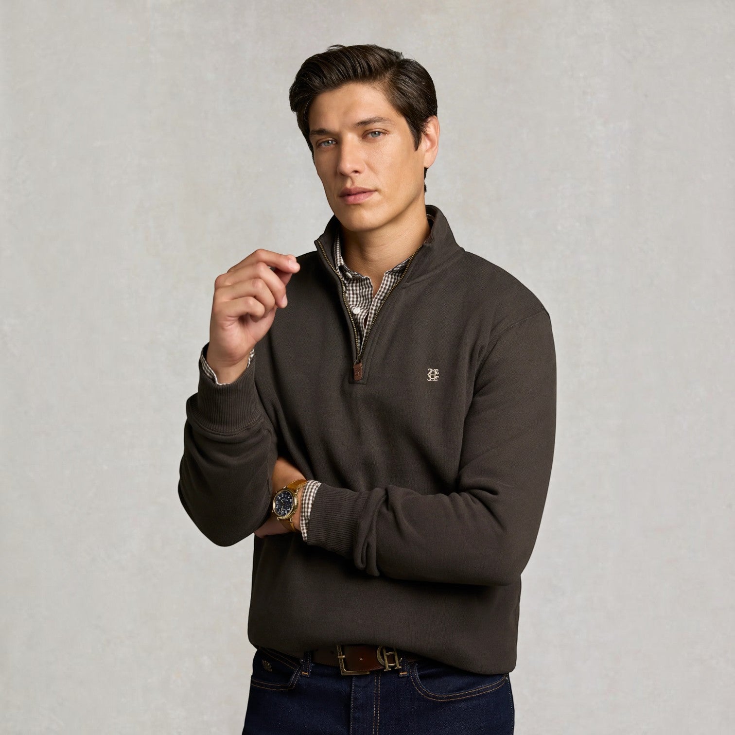 A man wearing a dark brown quarter-zip pullover sweater with a small embroidered logo on the chest, layered over a patterned collared shirt and paired with dark blue jeans, set against a textured light gray background.
