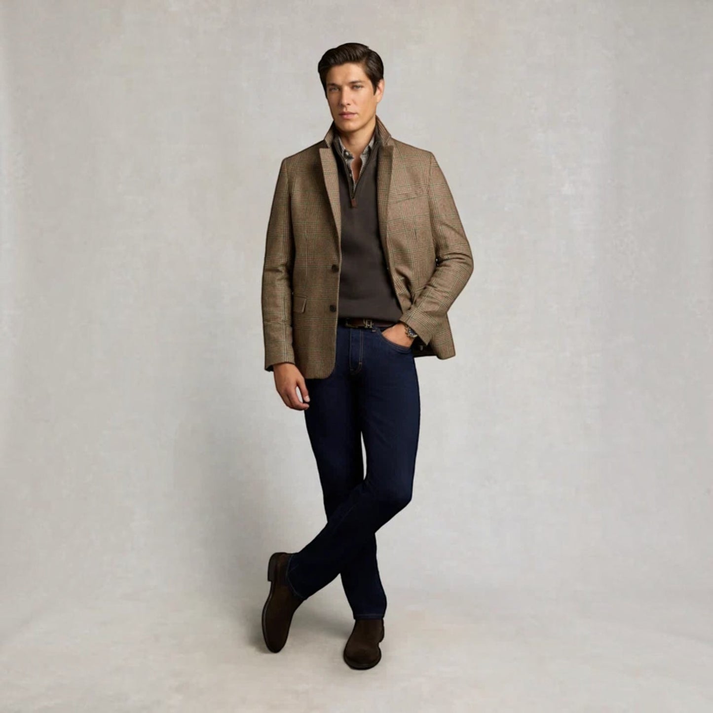 A man modeling a brown tweed blazer over a dark polo shirt, paired with dark blue jeans and brown suede boots, standing against a textured light gray studio backdrop.