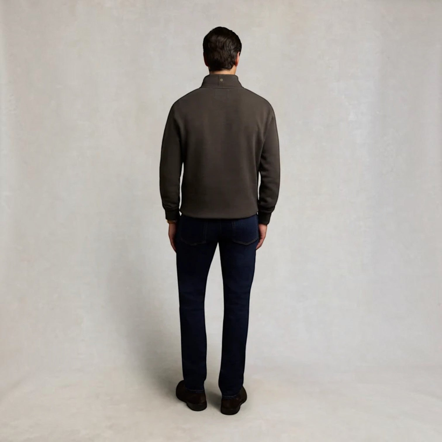 A man viewed from behind wearing a dark brown quarter-zip fleece pullover and dark blue jeans, standing against a neutral light gray textured studio backdrop.