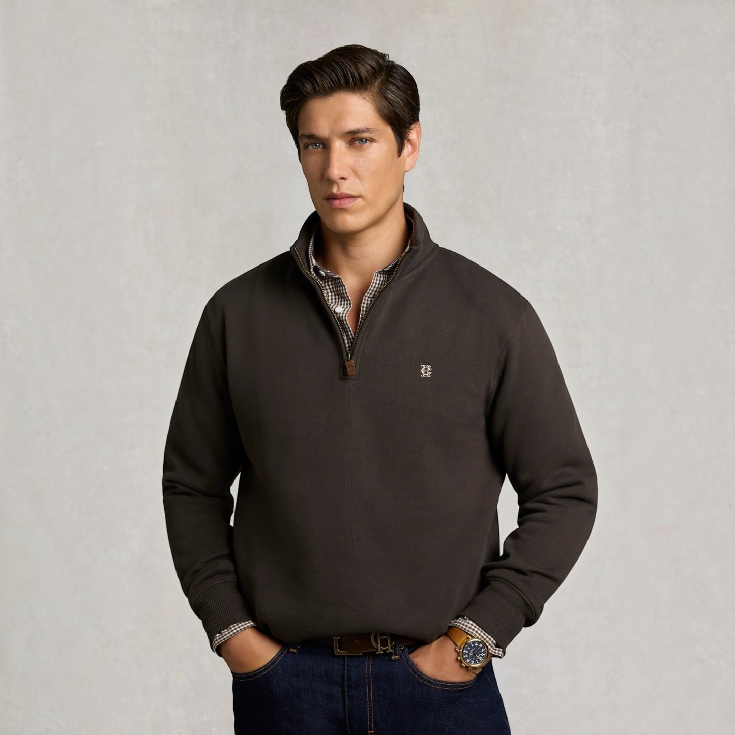 A man wearing a dark brown quarter-zip pullover sweater with a small embroidered logo on the chest, layered over a patterned collared shirt and paired with dark blue jeans, standing against a textured light gray background.