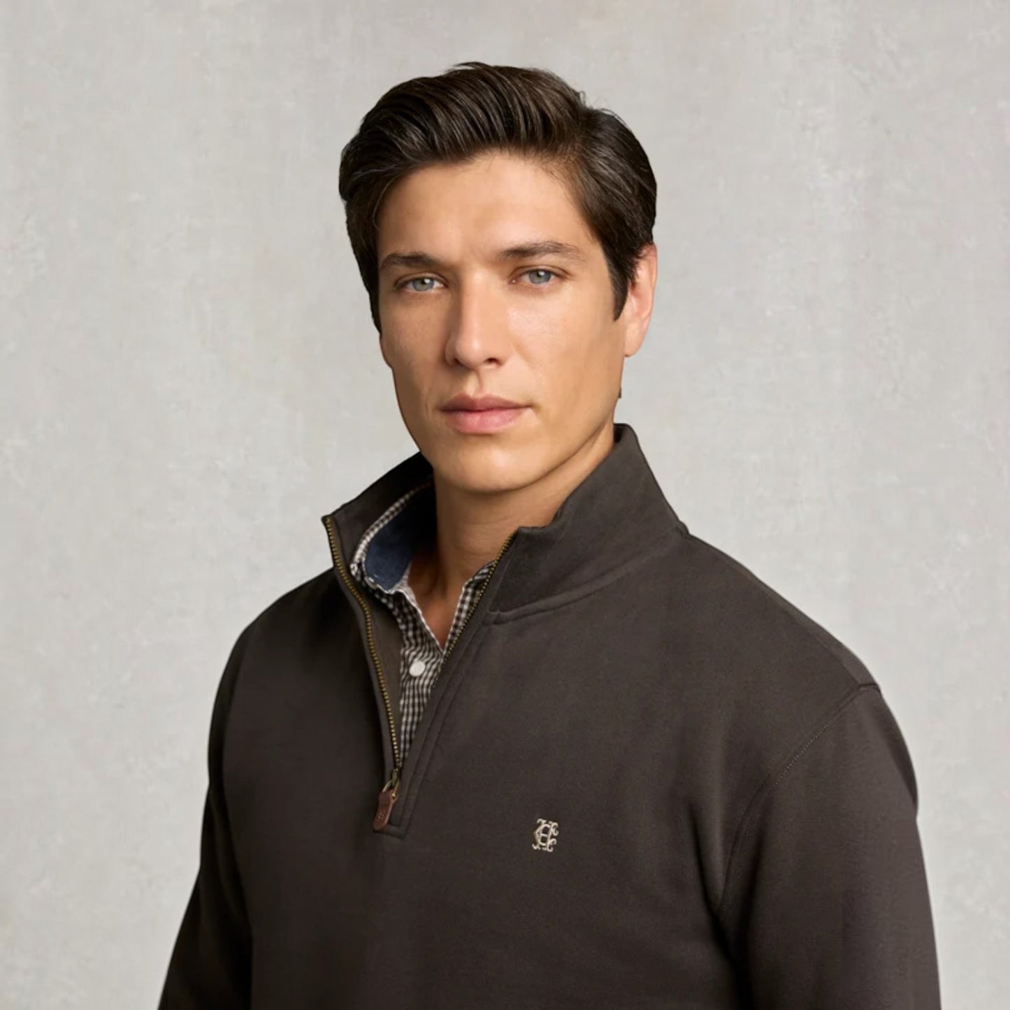 A man wearing a dark brown quarter-zip pullover sweater with a small embroidered logo on the chest, layered over a patterned collared shirt, set against a textured light gray background.