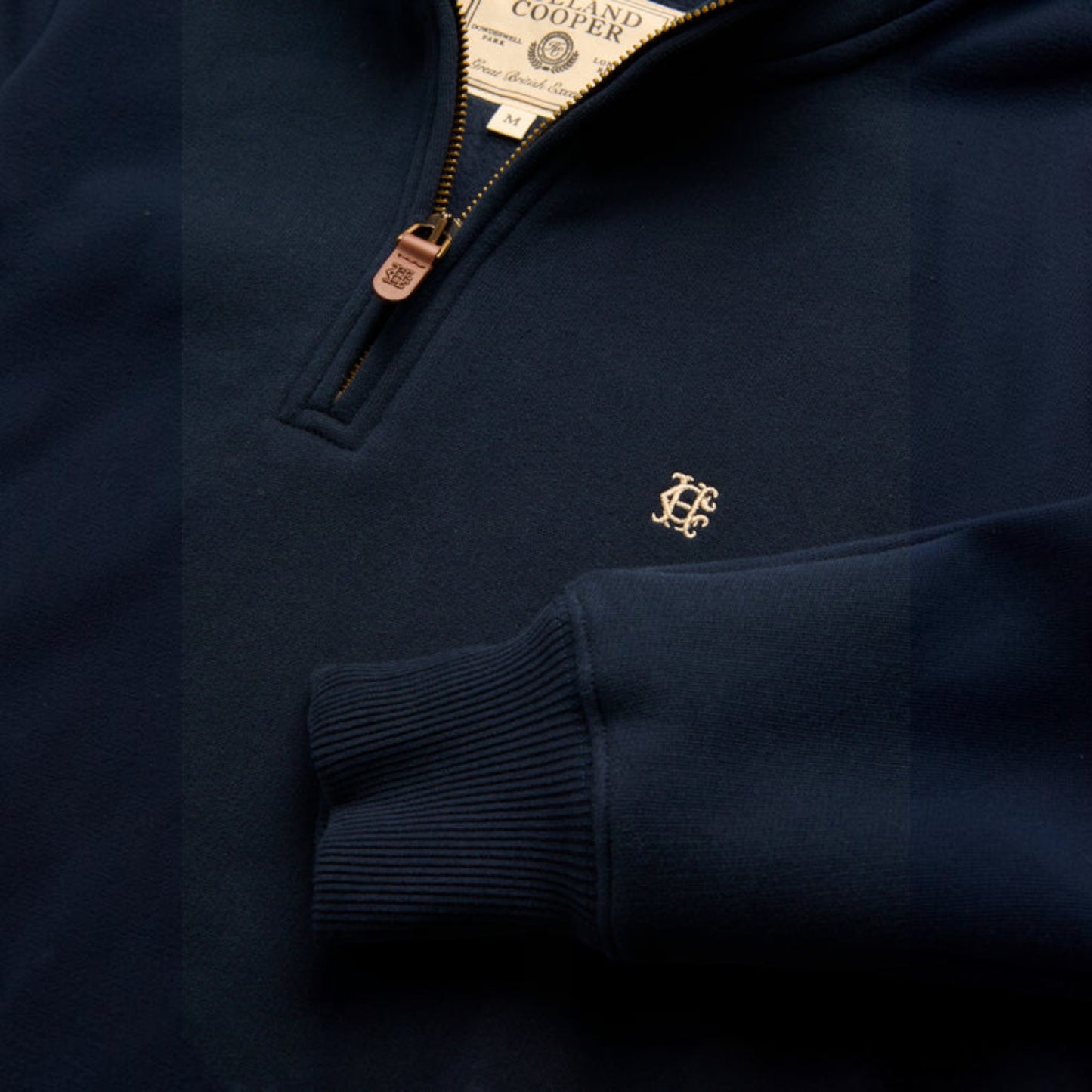Close-up of a navy blue quarter-zip sweatshirt with a small gold embroidered monogram logo on the chest and a visible neck label reading Cooper.