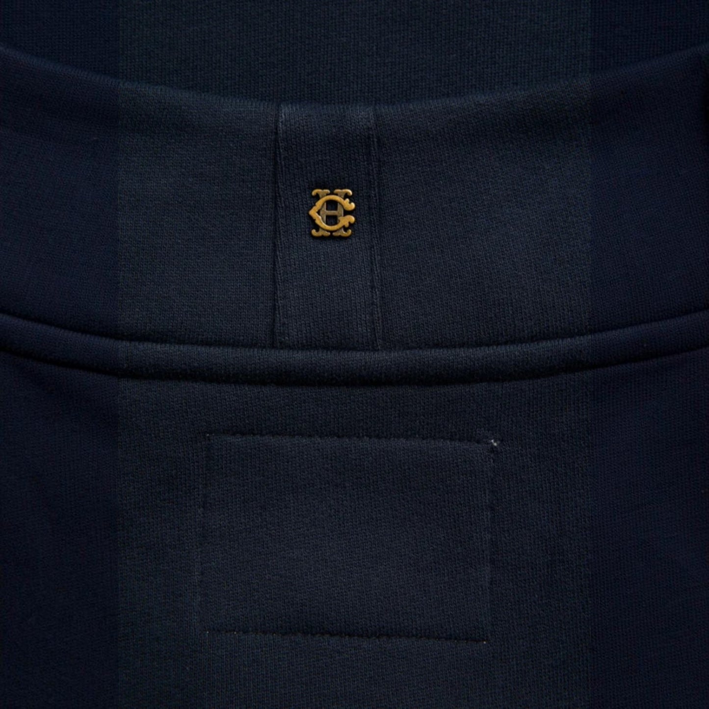 Close-up of a dark navy knit garment with a small gold embroidered circular logo centered below a waistband seam.