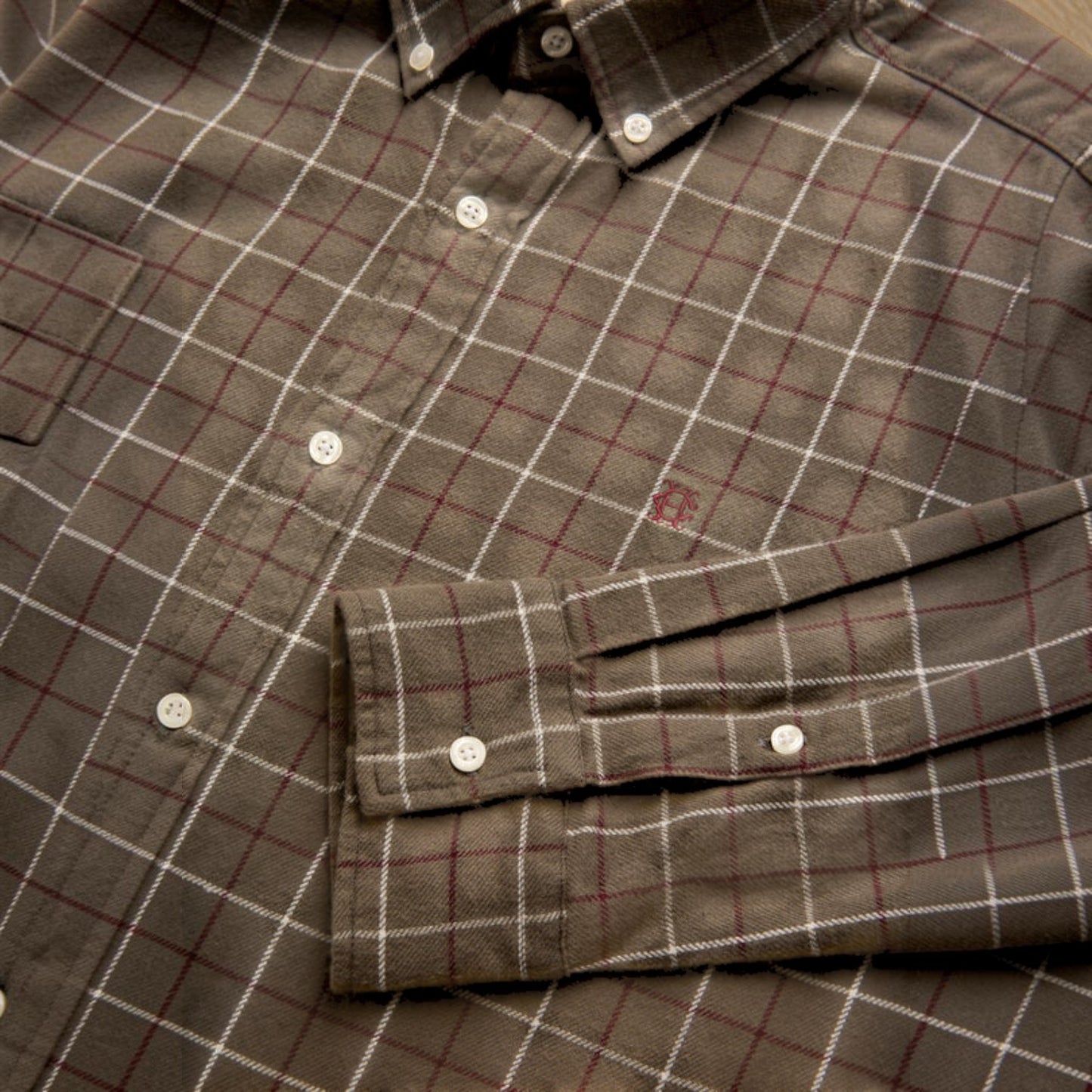 A close-up of a brown plaid button-down shirt with white and red grid lines, featuring a chest pocket and a folded cuff with snap buttons.
