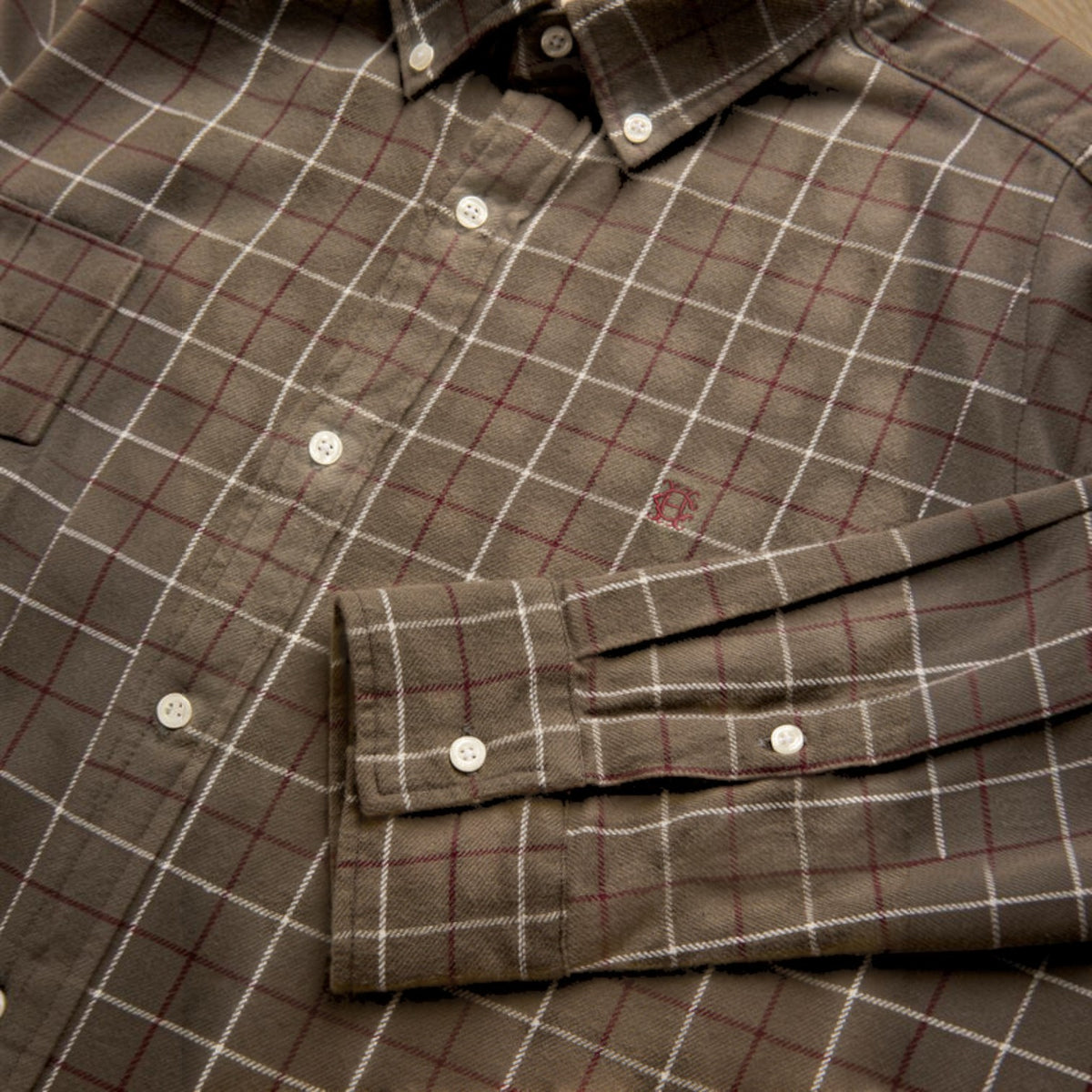 A close-up of a brown plaid button-down shirt with white and red grid lines, featuring a chest pocket and a folded cuff with snap buttons.