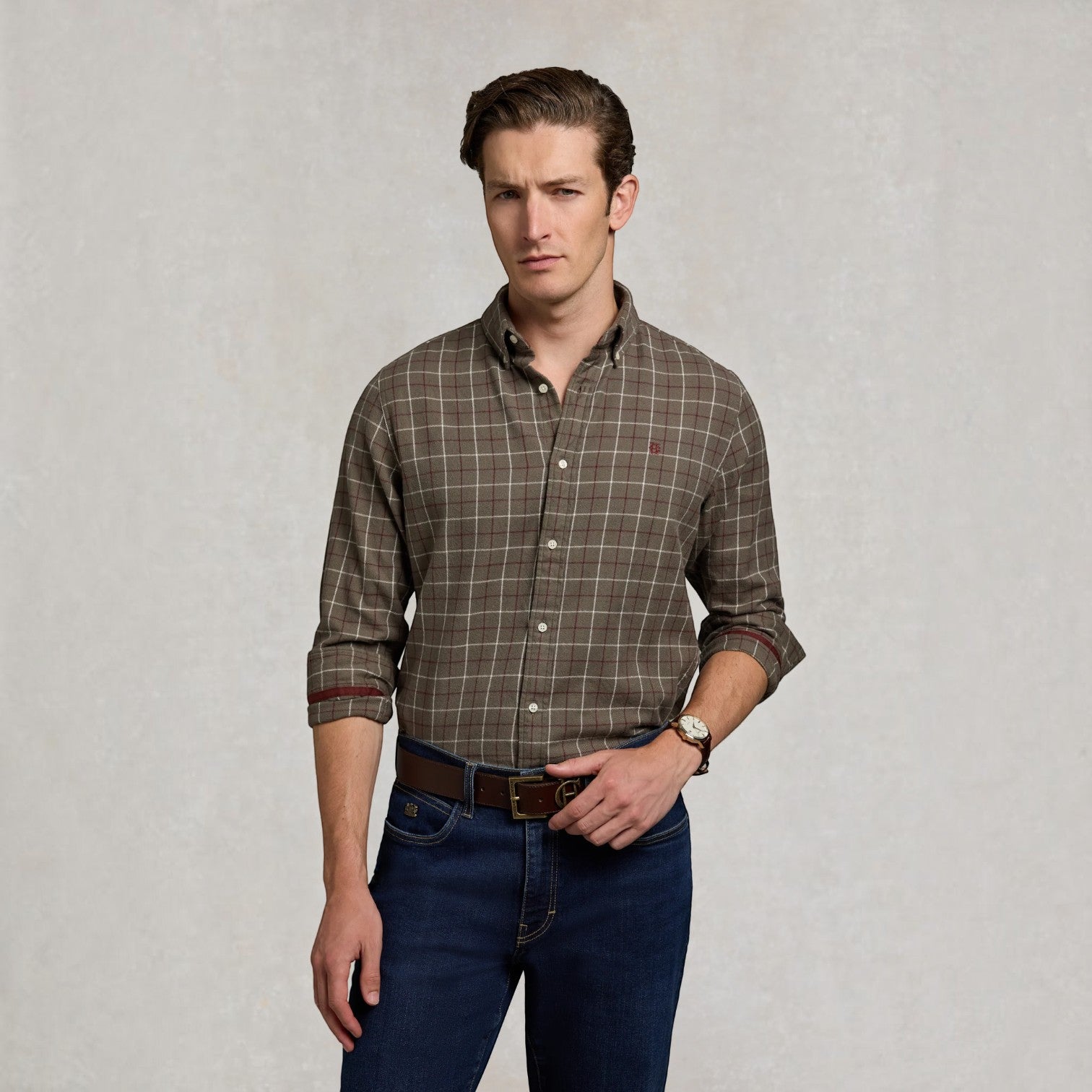 A man wearing a brown and beige plaid button-down shirt with rolled-up sleeves, paired with dark blue jeans and a brown belt, standing against a textured light gray background.