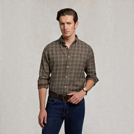 A man wearing a brown and beige plaid button-down shirt with rolled-up sleeves, paired with dark blue jeans and a brown belt, standing against a textured light gray background.