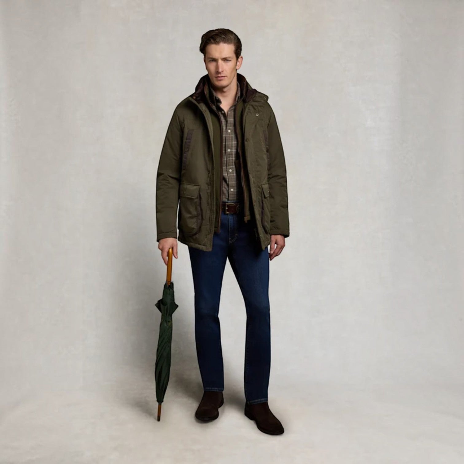 A man stands against a light gray studio background wearing an olive green hooded jacket over a plaid shirt and blue jeans, holding a dark green umbrella.
