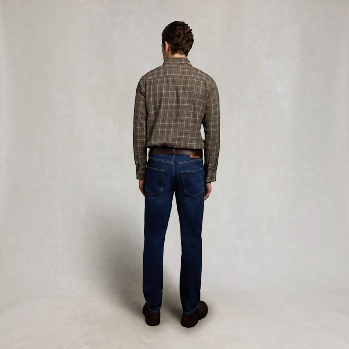 A man viewed from behind wearing a brown and beige plaid button-down shirt, dark blue jeans, and brown shoes, standing against a neutral light gray studio background.
