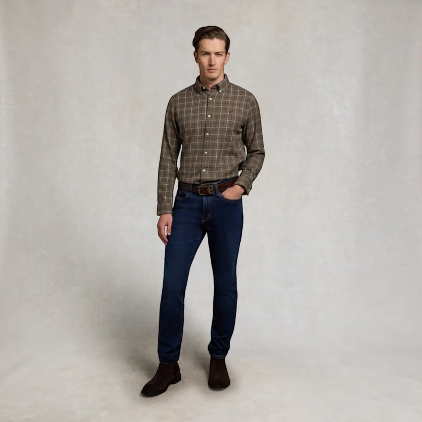 A man modeling a brown and beige plaid long-sleeve button-down shirt, paired with dark blue jeans, a brown belt, and dark brown boots, standing against a neutral textured studio backdrop.