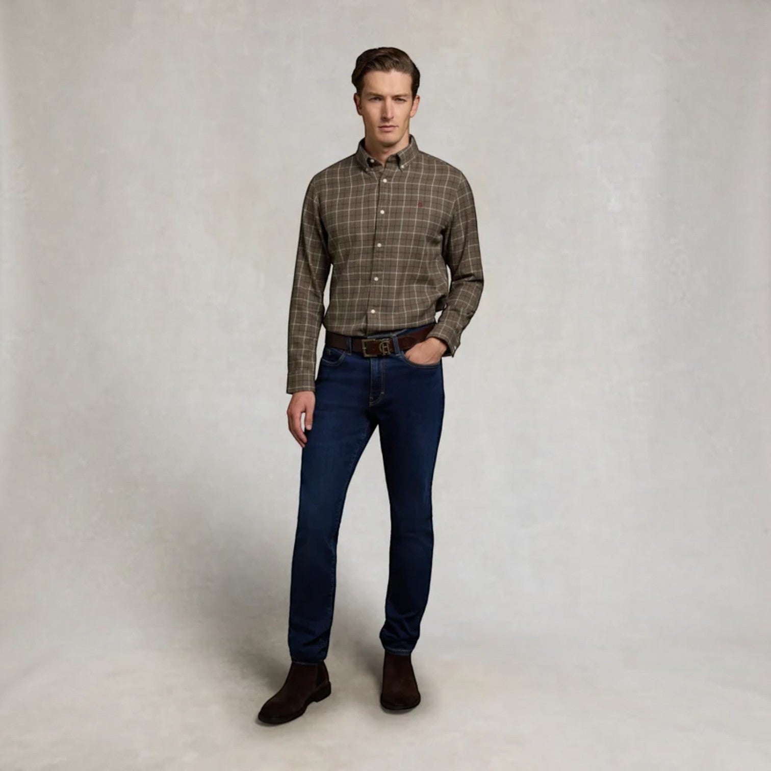 A man modeling a brown and beige plaid long-sleeve button-down shirt, paired with dark blue jeans, a brown belt, and dark brown boots, standing against a neutral textured studio backdrop.