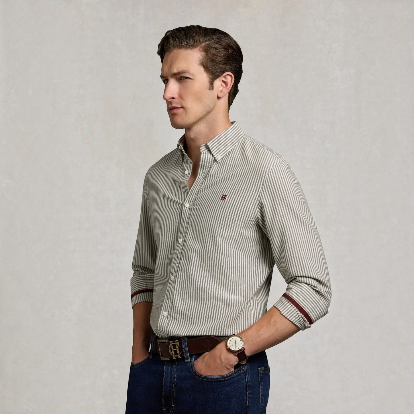 A man wearing a slim-fit, long-sleeve button-down shirt with fine vertical stripes in beige and dark brown, paired with blue jeans and a brown leather belt, standing against a textured light gray background.