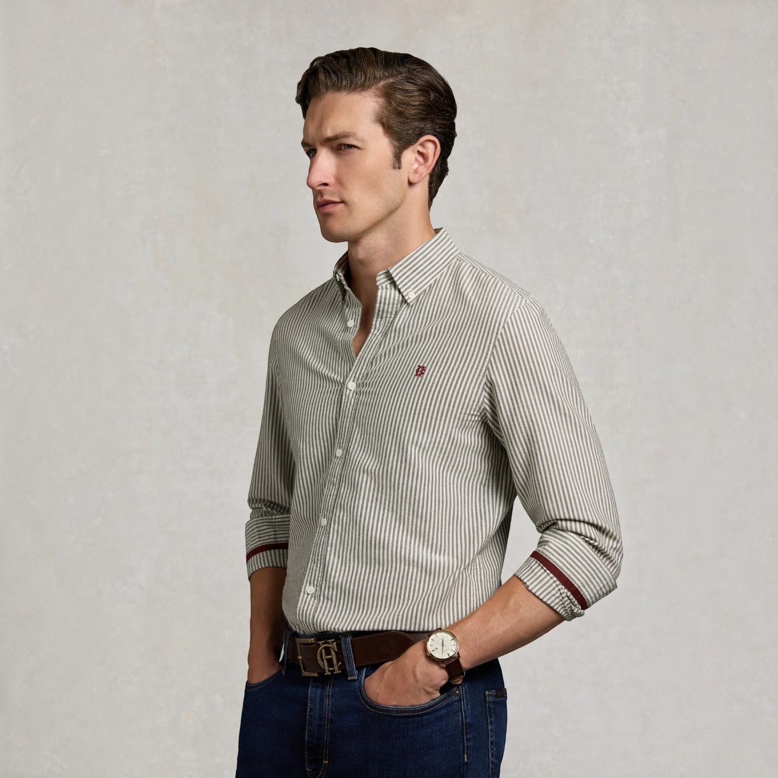 A man wearing a slim-fit, long-sleeve button-down shirt with fine vertical stripes in beige and dark brown, paired with blue jeans and a brown leather belt, standing against a textured light gray background.