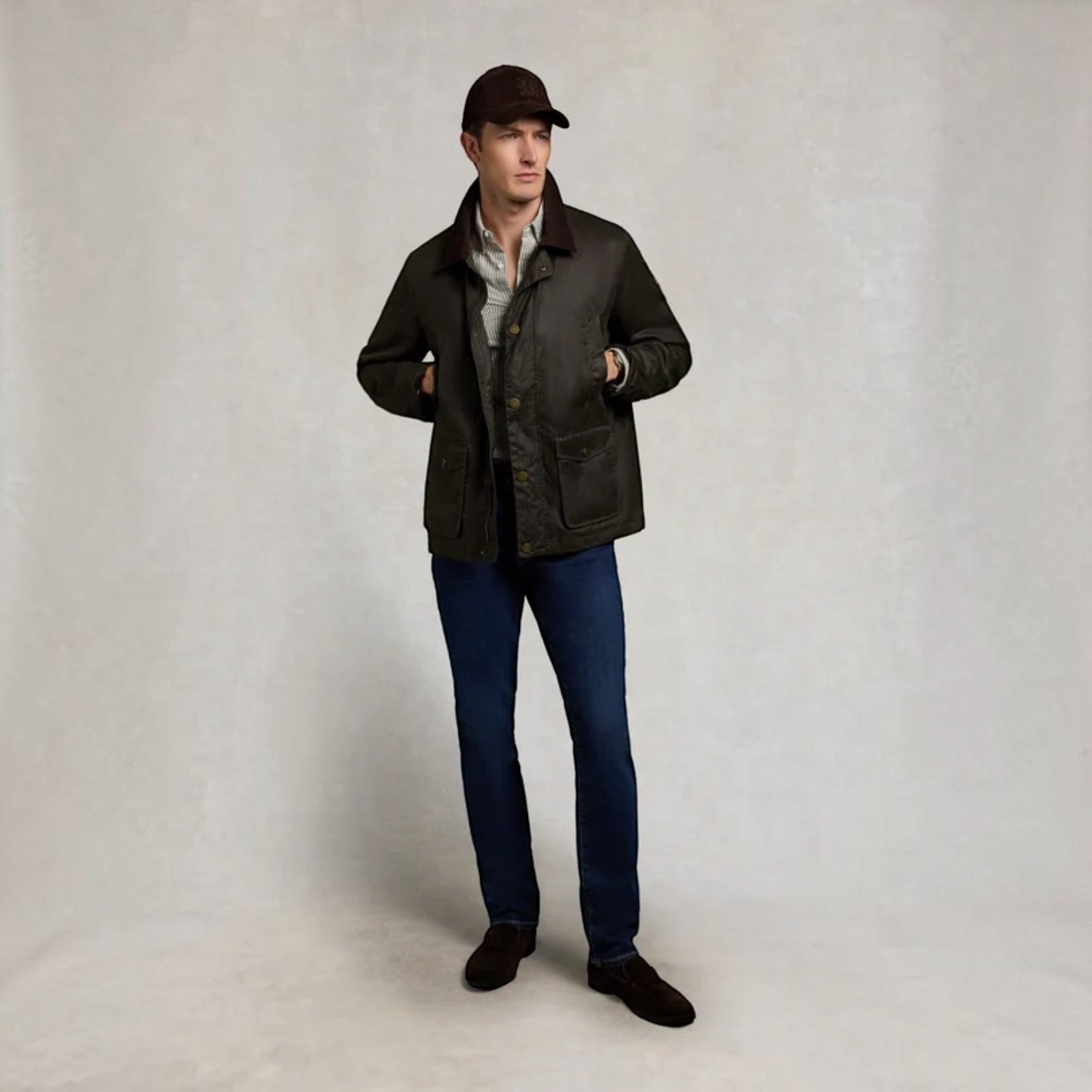 A man modeling a dark brown waxed canvas jacket, blue jeans, a light-colored shirt, and a dark brown cap, standing against a neutral textured studio backdrop.