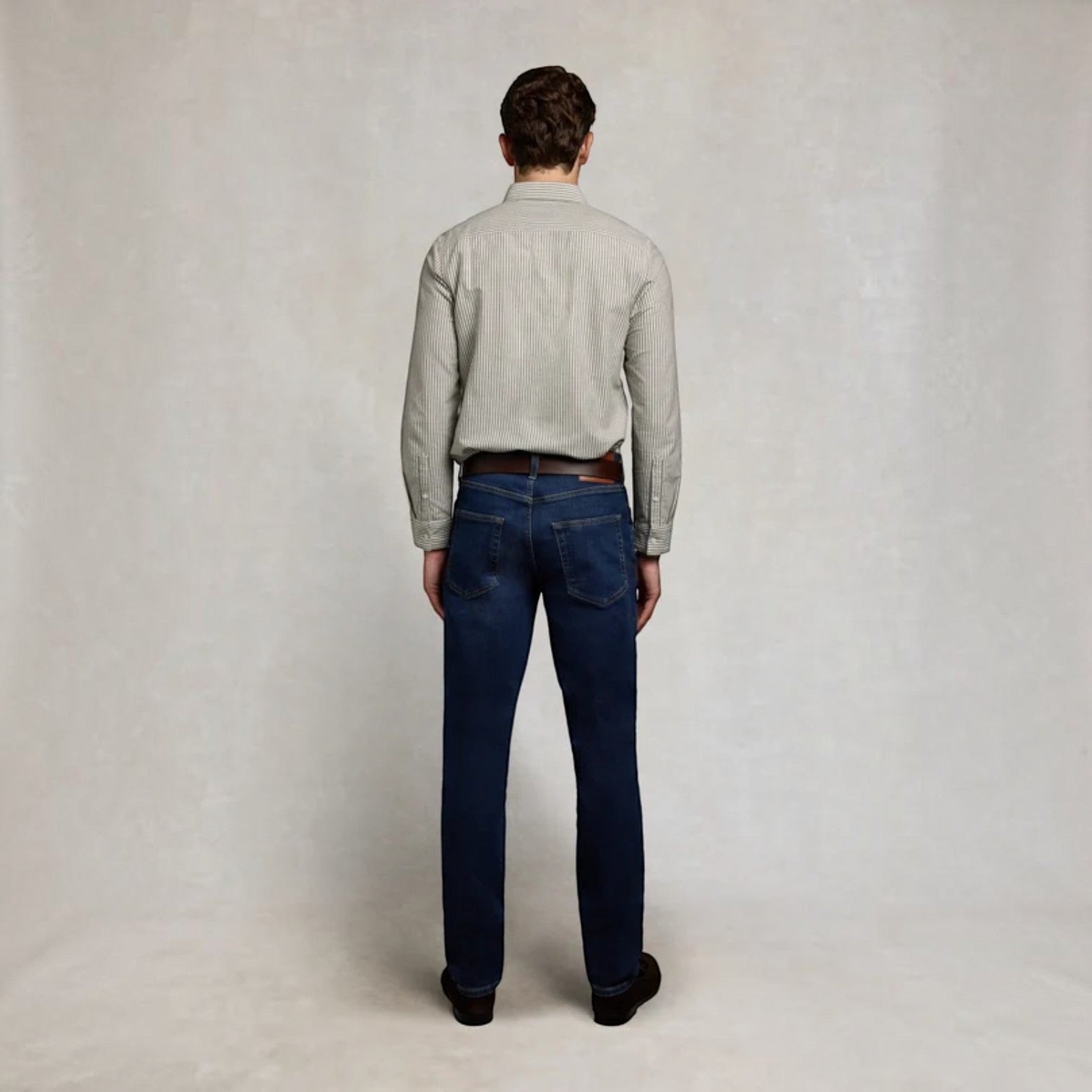 A man viewed from behind wearing a light gray long-sleeve button-down shirt and dark blue jeans, standing against a neutral light gray studio backdrop.