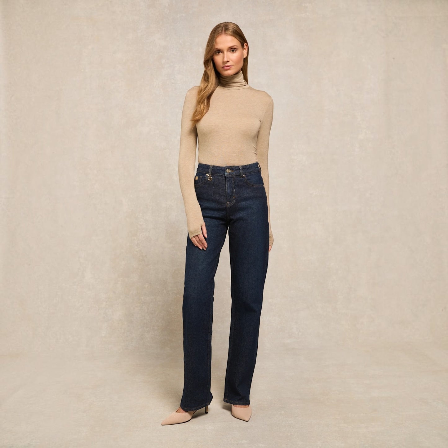 A woman stands against a textured beige studio backdrop wearing a beige turtleneck sweater, dark blue straight-leg jeans, and beige pointed-toe heels.